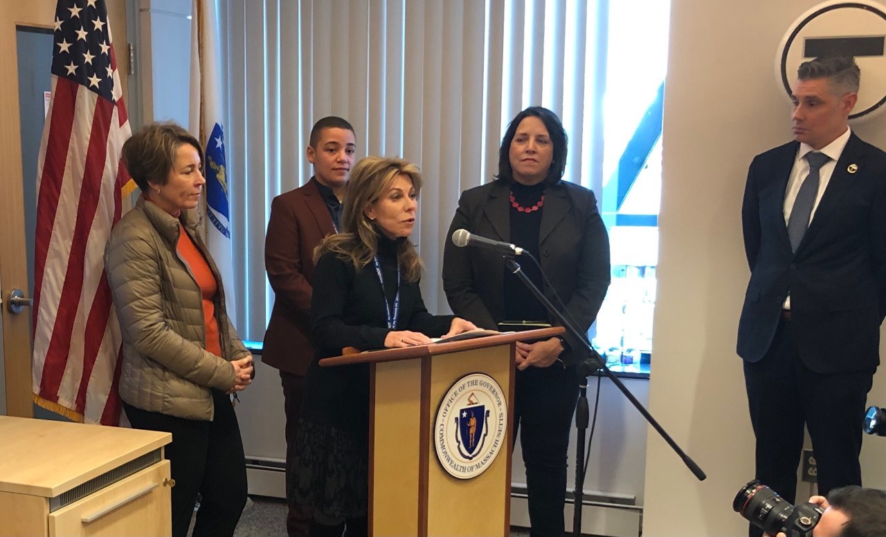 Four women in suits stand around a podium with a microphone and the seal of Massachusetts. An American flag is behind them. A man in a suit and tie stands to the side in front of an MBTA "T" logo. 