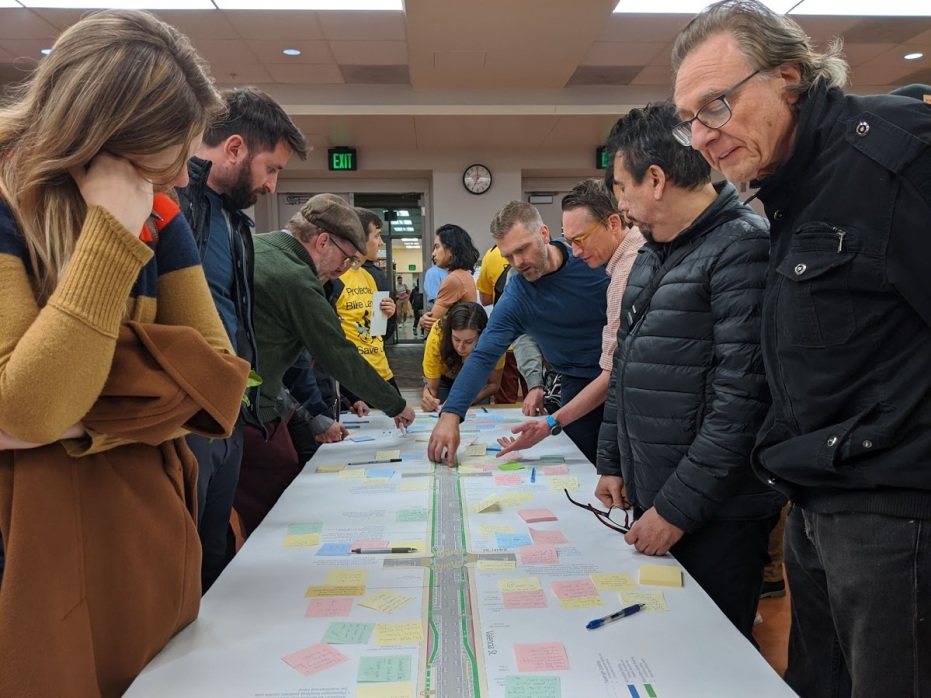 A crowd of people gathers around a table covered in the layout plan for a street. Some people are placing sticky notes on the diagram.