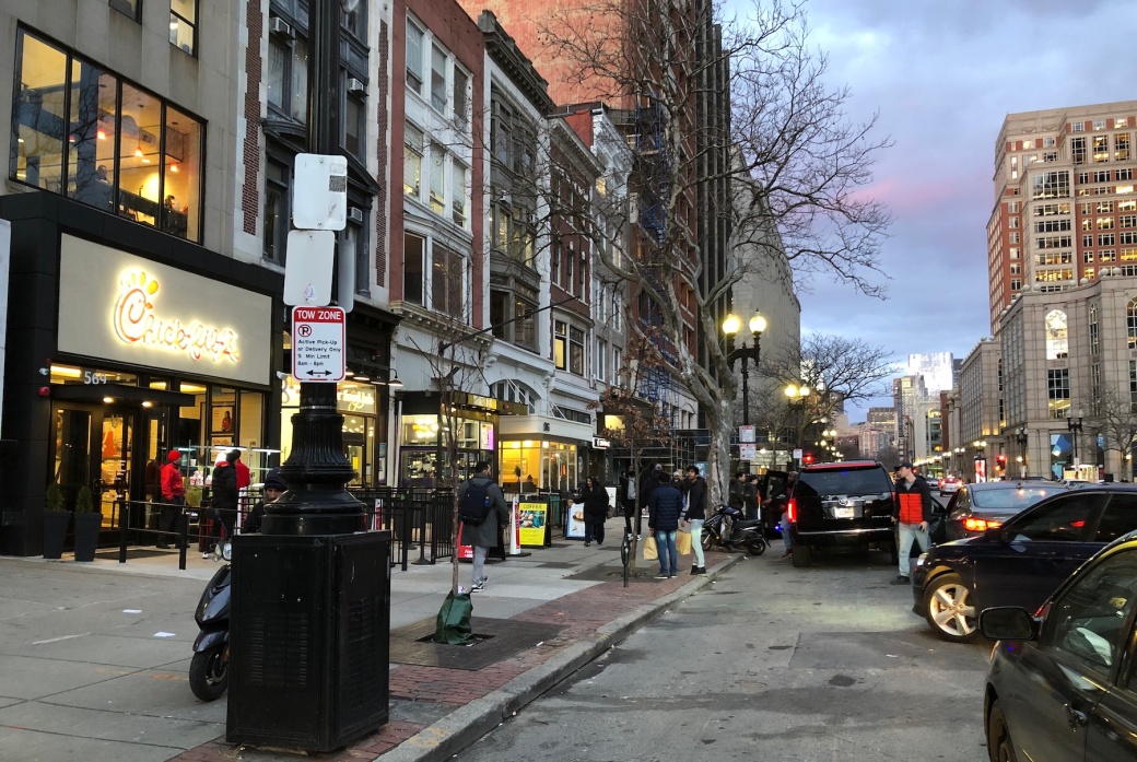 An evening city street scene on Boylston Street near Copley Square. On the left side of the photo is a busy sidewalk and a Chick-Fil-A restaurant. On the street in front of the restaurant, four cars, three of them double-parked, occupy the multiple-lane street; a food delivery driver is stepping out of one of the cars while a second car is attempting to drive between the others to fit in closer to the curb.