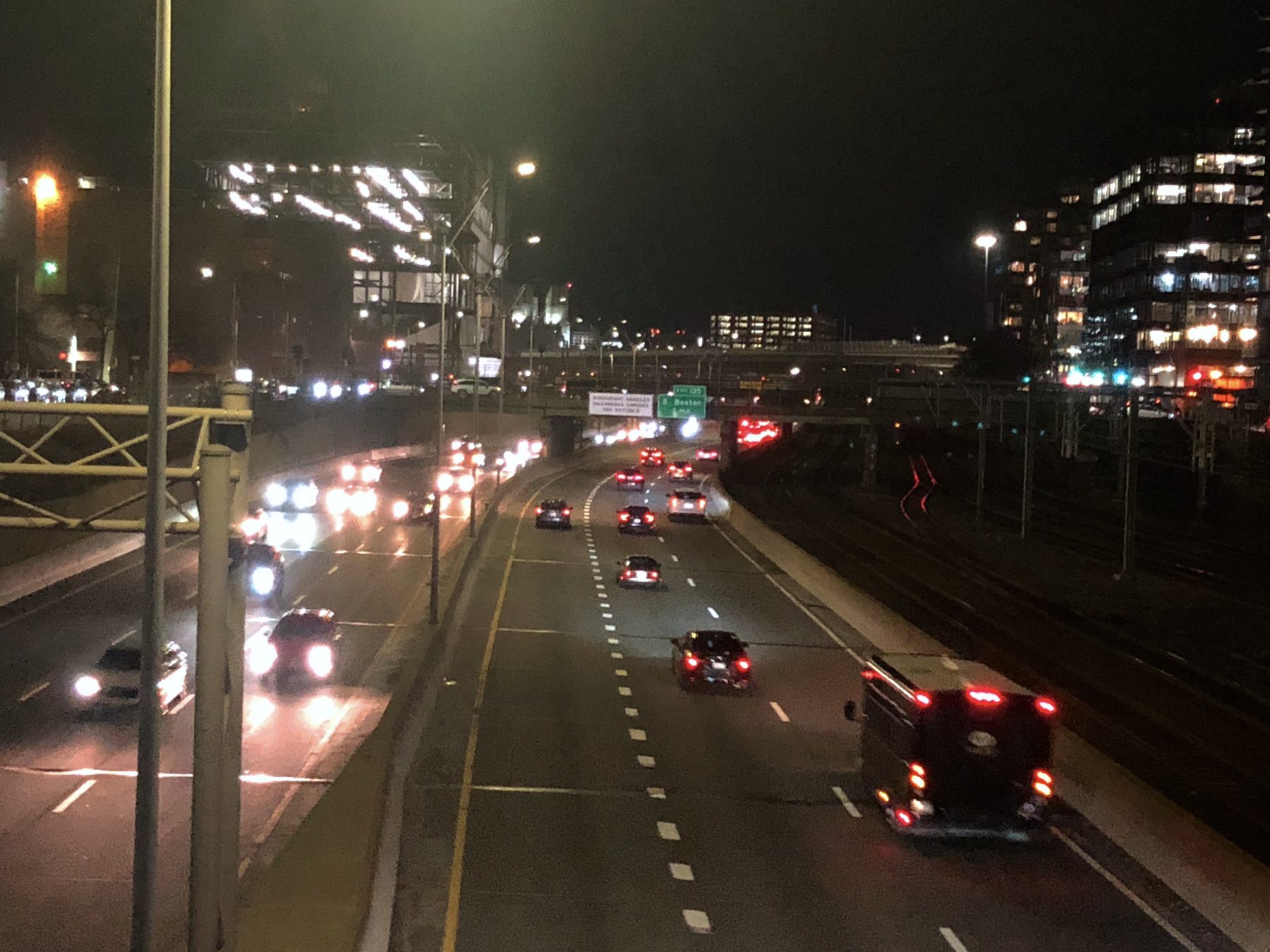 A nighttime view of cars and trucks on a 6-lane expressway next to some railroad tracks (right) in downtown Boston.