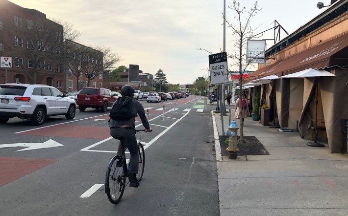 A bicyclist rides in a bike lane alongside a row of flexible-post bollards that divide the bike lane from a maroon-painted bus lane. A sign on the curb says "RIGHT LANE BUSES ONLY". An awning over the adjacent sidewalk covers an outdoor dining area in front of a restaurant.
