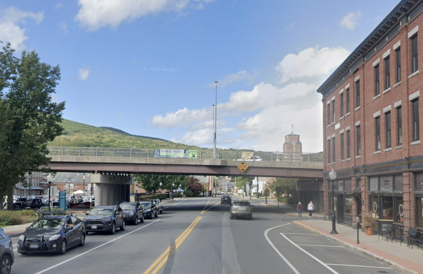 A view of a downtown street lined withparked cars and historic 3-story brick buildings with a highway overpass crossing over the street in the middle distance. In the background are green hills below a blue sky.