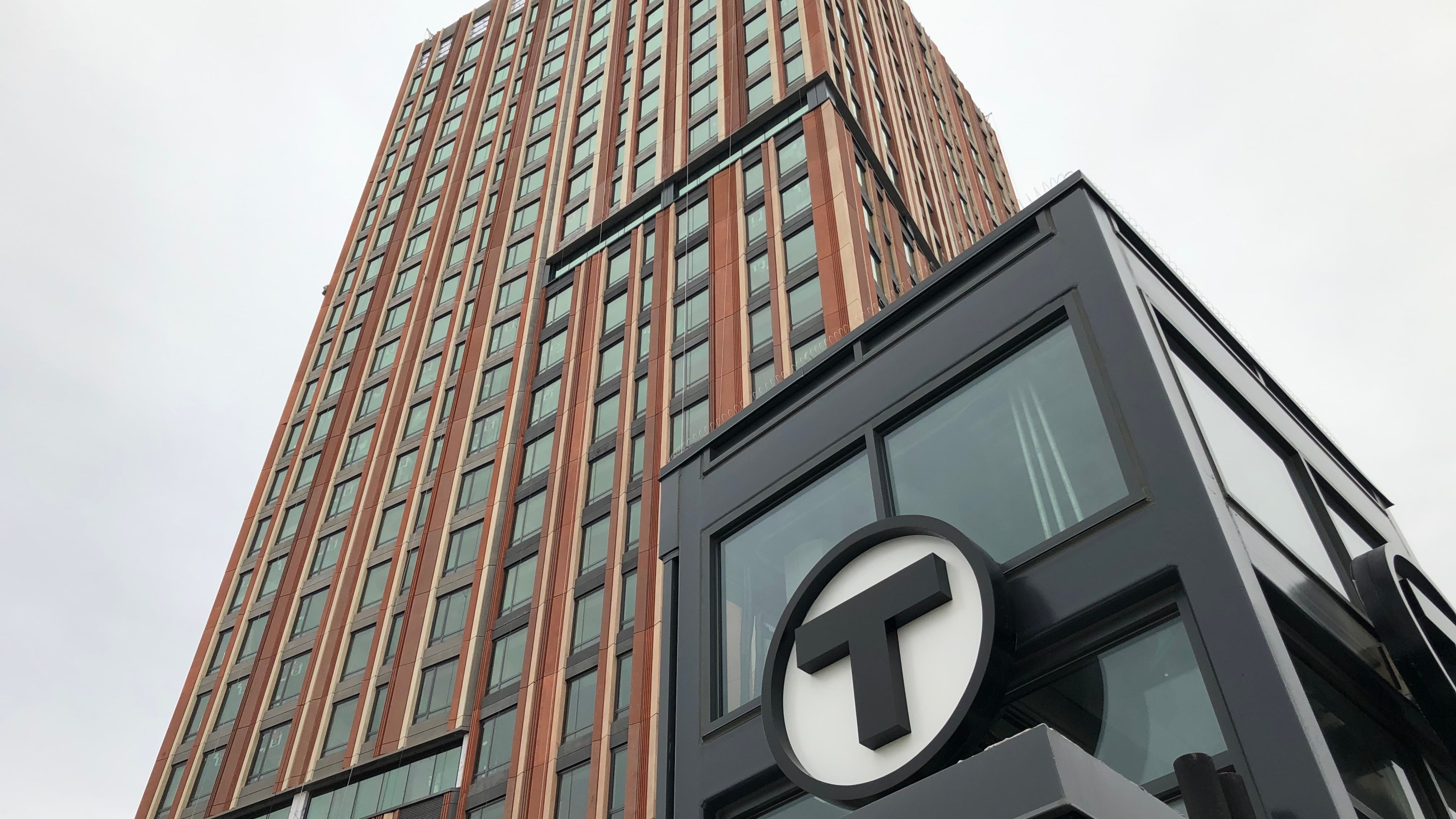 An apartment tower under construction, seen from the ground angle looking up into the sky. In the foreground is a black elevator enclosure with the MBTA T logo.