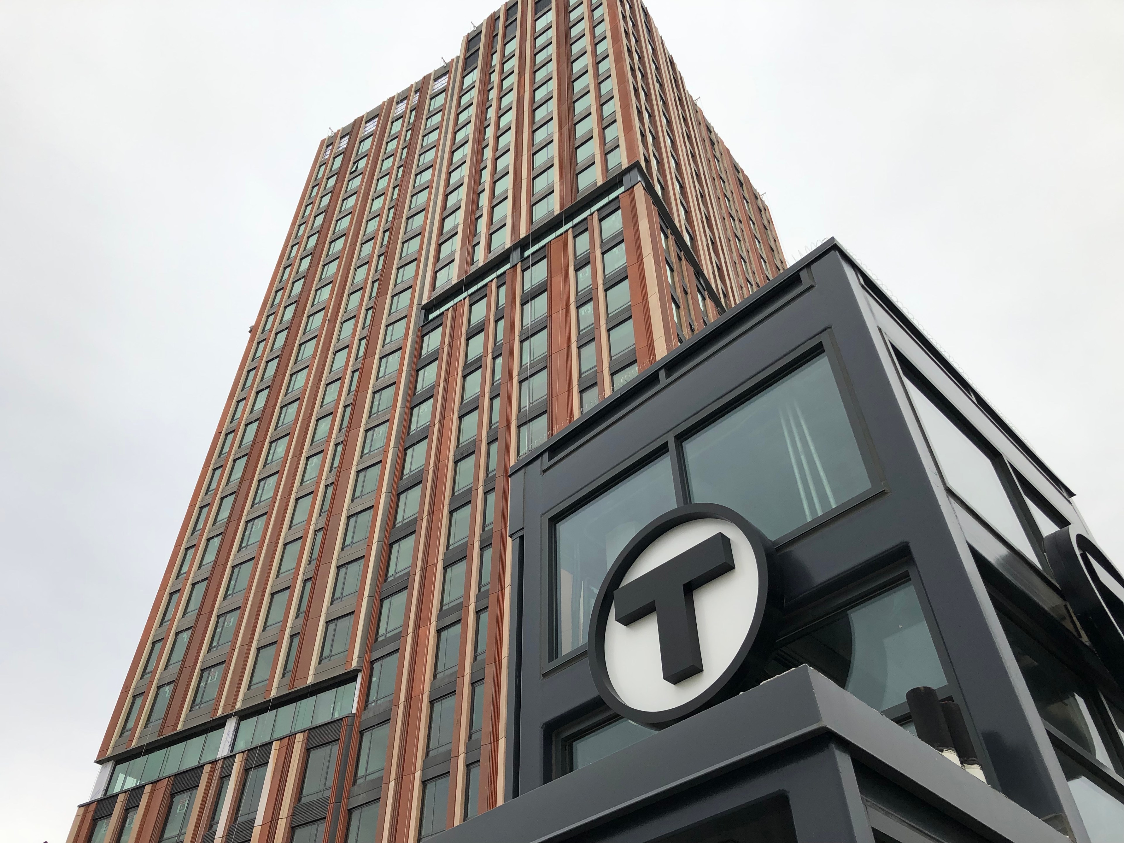 An apartment tower under construction, seen from the ground angle looking up into the sky. In the foreground is a black elevator enclosure with the MBTA T logo.