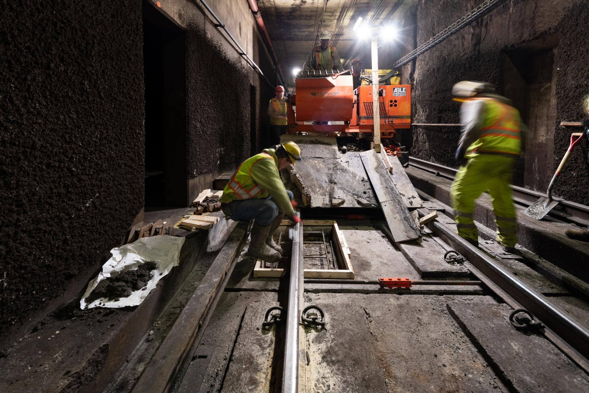 Workers wearing flourescent green jackets place new rebar under the subway tracks inside a subway tunnel. Behind them, three more workers watch over them from the top of an orange machine that has lights illuminating the work