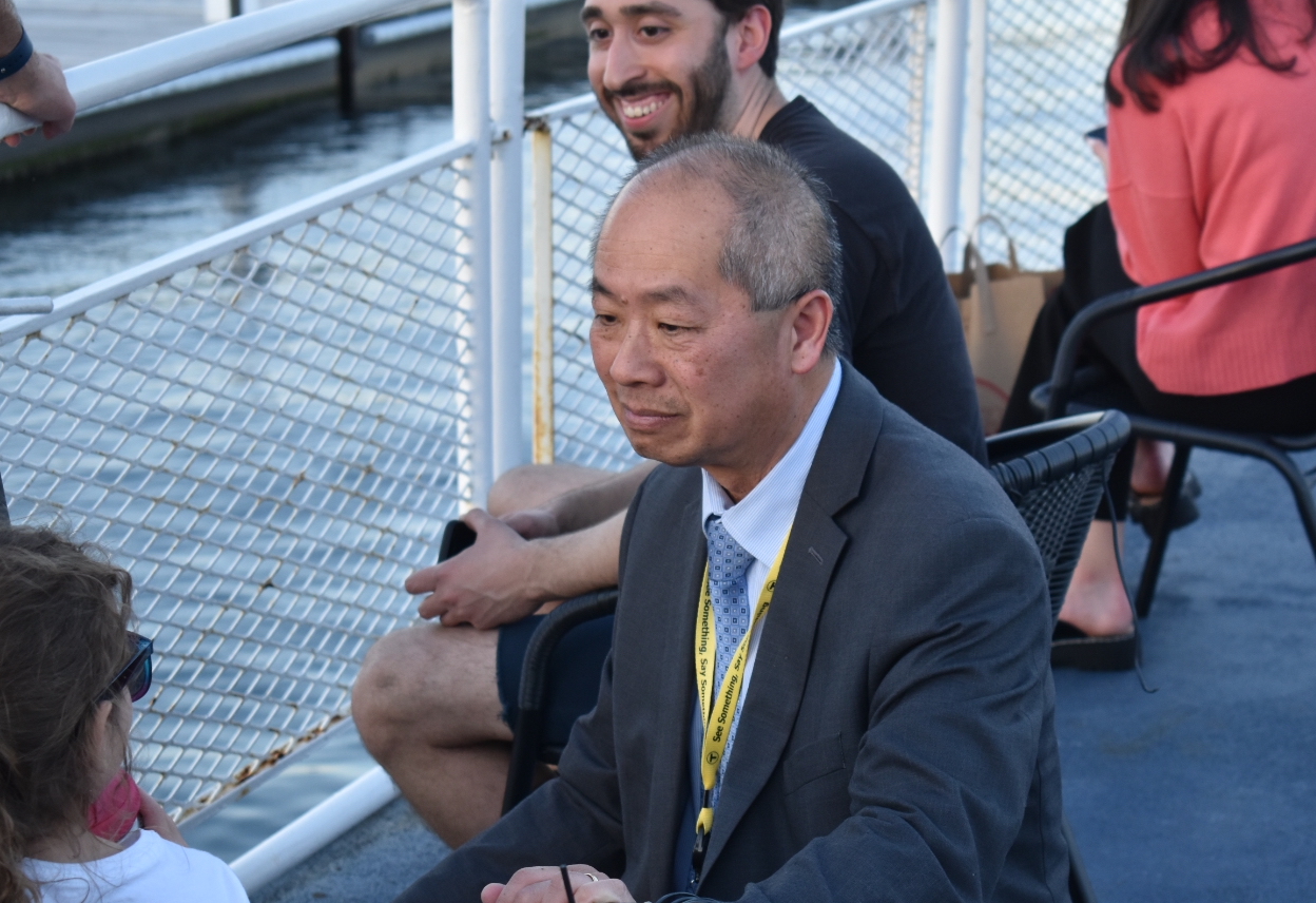 A middle-aged Asian man in a suit and tie squats down to talk with a child aboard a ferry boat. An adult looks on behind him, and beyond the boat's railing the water is visible.