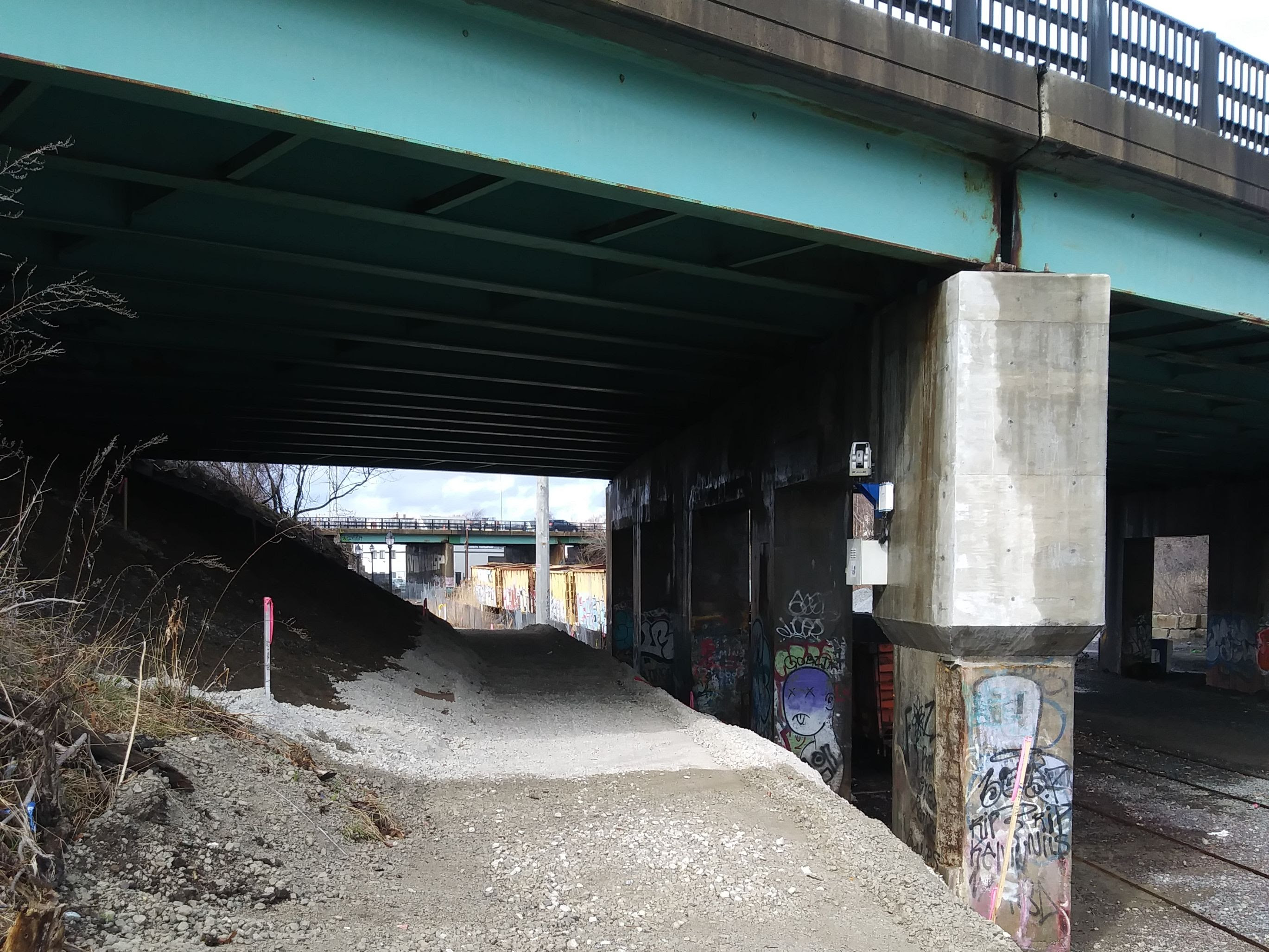 A graded gravel path under construction runs underneath a highway bridge next to a concrete bridge abutment. To the right of the abutment and below the trail are some railroad tracks.