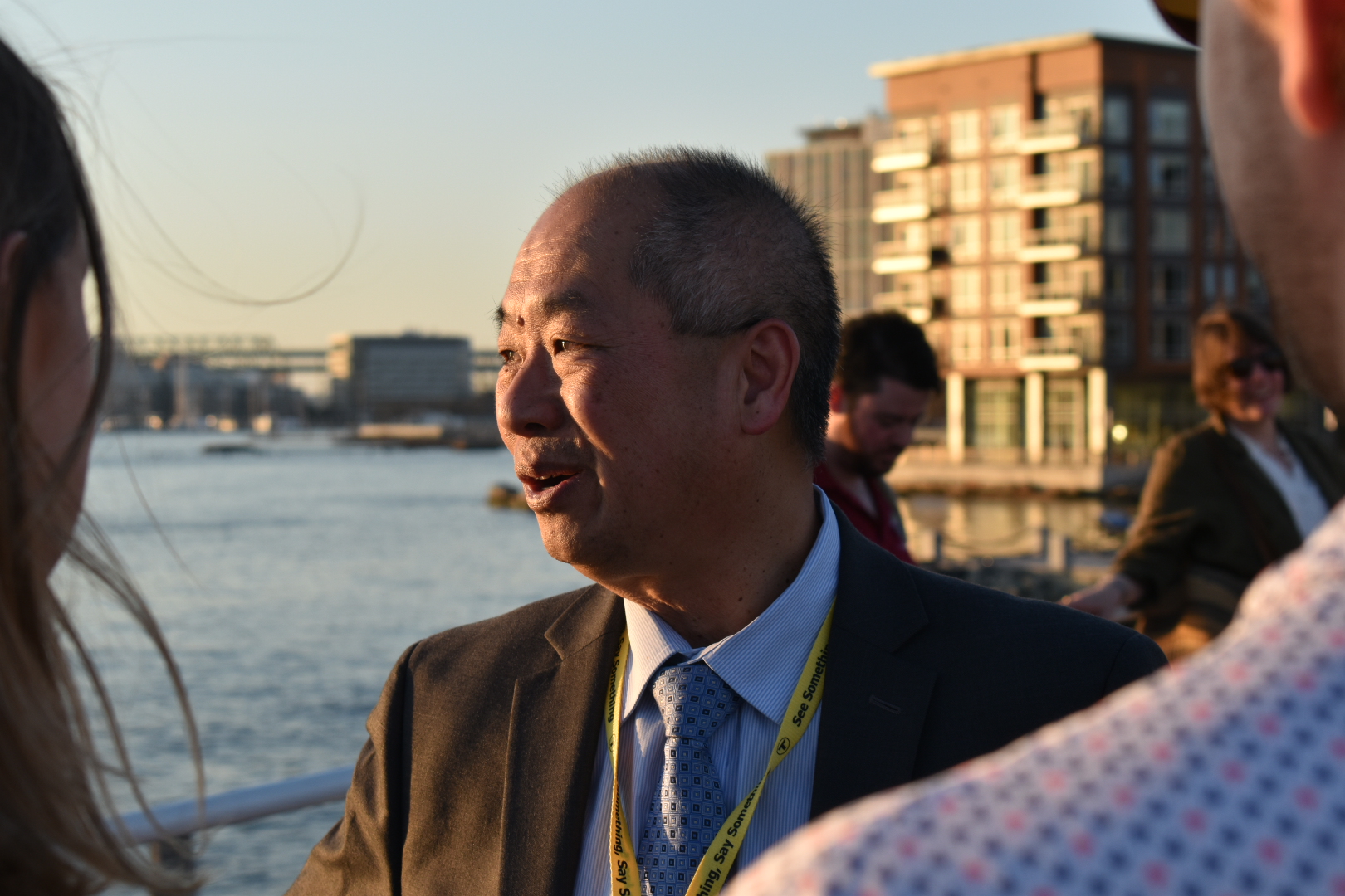 A middle-aged Asian man in a suit and tie looks into the setting sun with the Boston Harbor behind him.