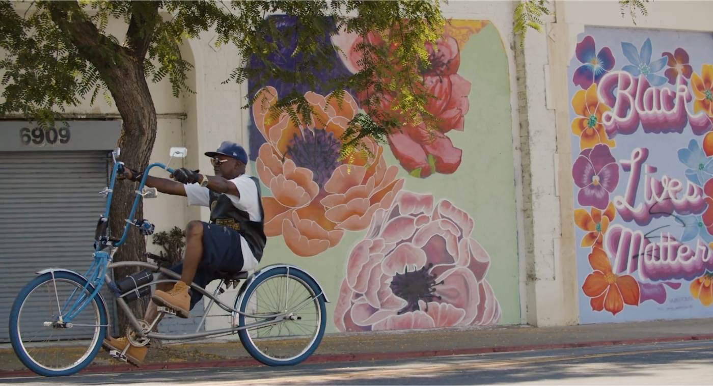 A Black man wearing shorts and a t-shirt rides a chopper bicycle past a mural with flowers that says "Black Lives Matter"