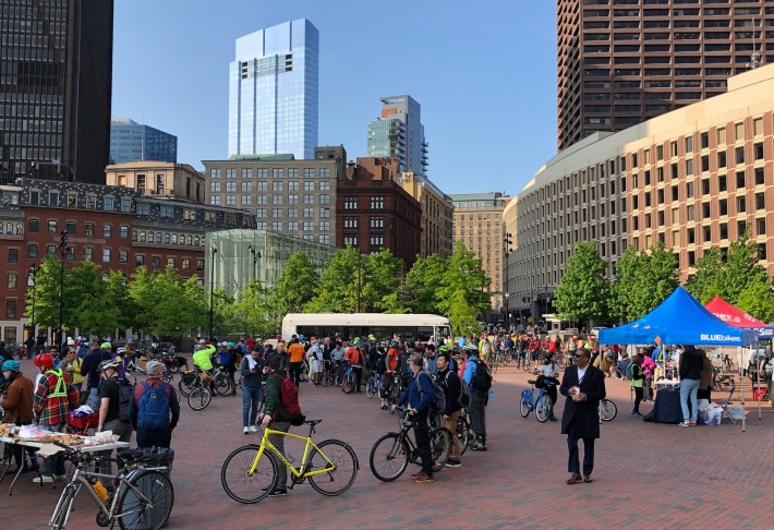 A crowd of over 100 people, many of whom are walking with their bicycles, gathers in a large brick-paved plaza. In the distance are the skyscrapers of downtown Boston.