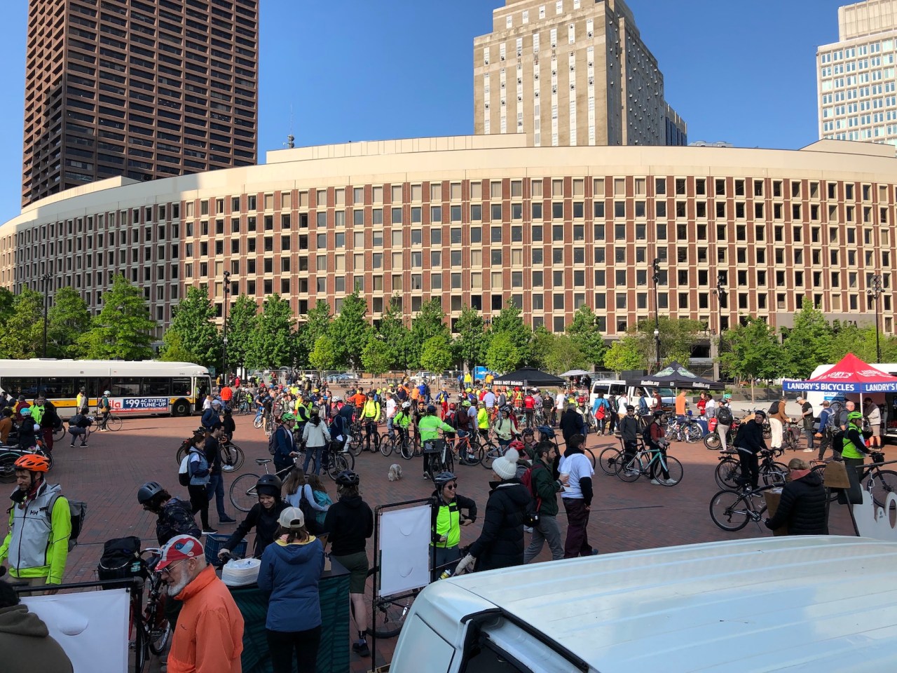 A crowd of over 100 people, many of whom are walking with their bicycles, gathers in a large brick-paved plaza. Several tents around the edge of the plaza advertise local bike shops. In the distance are the skyscrapers of downtown Boston.