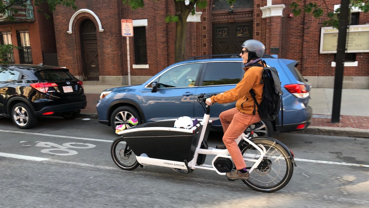 A man wearing salmon-colored pants and an orange jacket pedals a white Dutch-style cargo bike along a bike lane next to a parked car. His young son, visible only from the white bike helmet he's wearing, is sitting in the cargo bucket in front of him.
