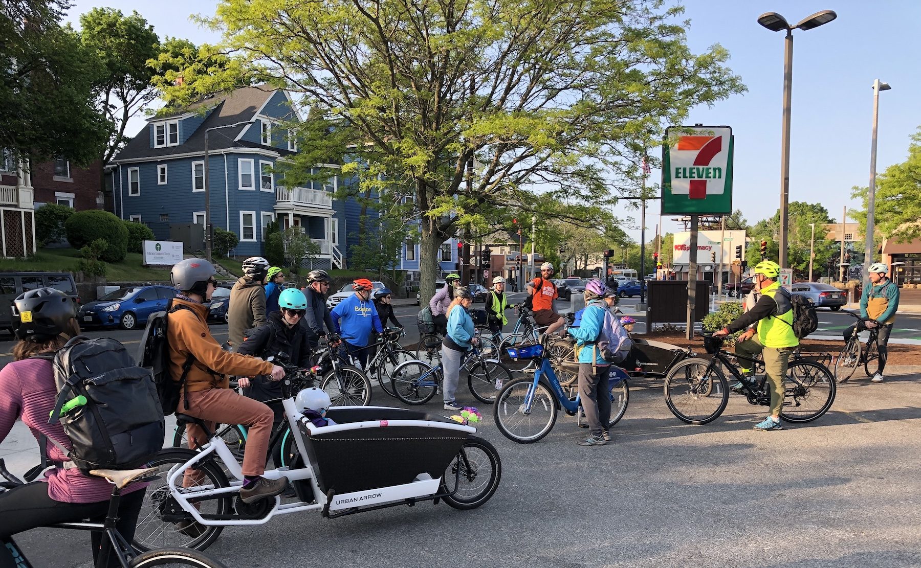 A crowd of bike riders gathers in a parking lot beneath a 7-11 sign. Several of the people are riding Dutch-style cargo bikes with large front-mounted boxes in which their children are sitting to come along for the ride.