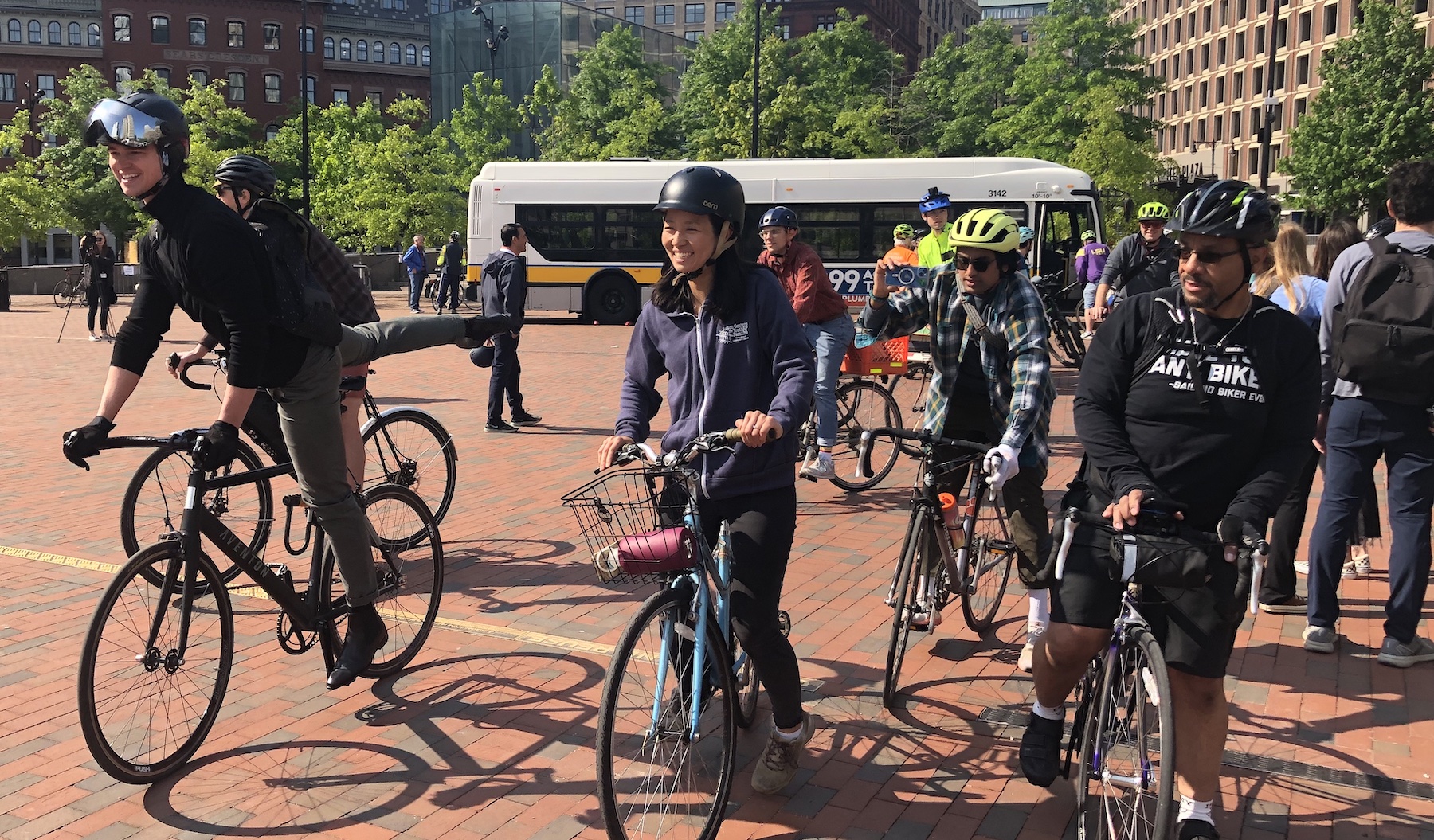 A crowd of people in bikes pedals through a wide brick plaza. In the center, wearing a black helmet and blue hoodie, is Boston Mayor Michelle Wu.