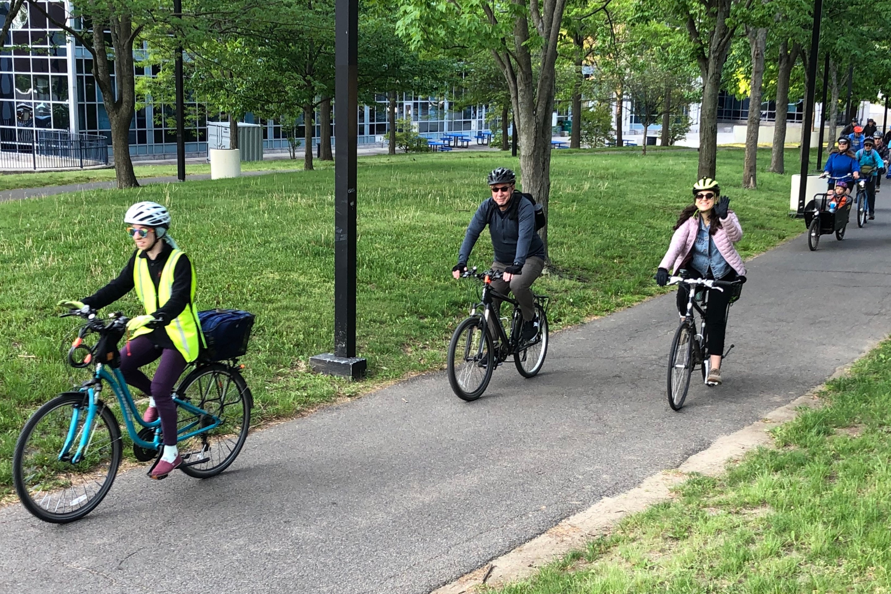 Three people riding bicycles down a paved pathway through a shady park on the SW Corridor. Behind them in the upper right corner is another crowd of people on bikes.