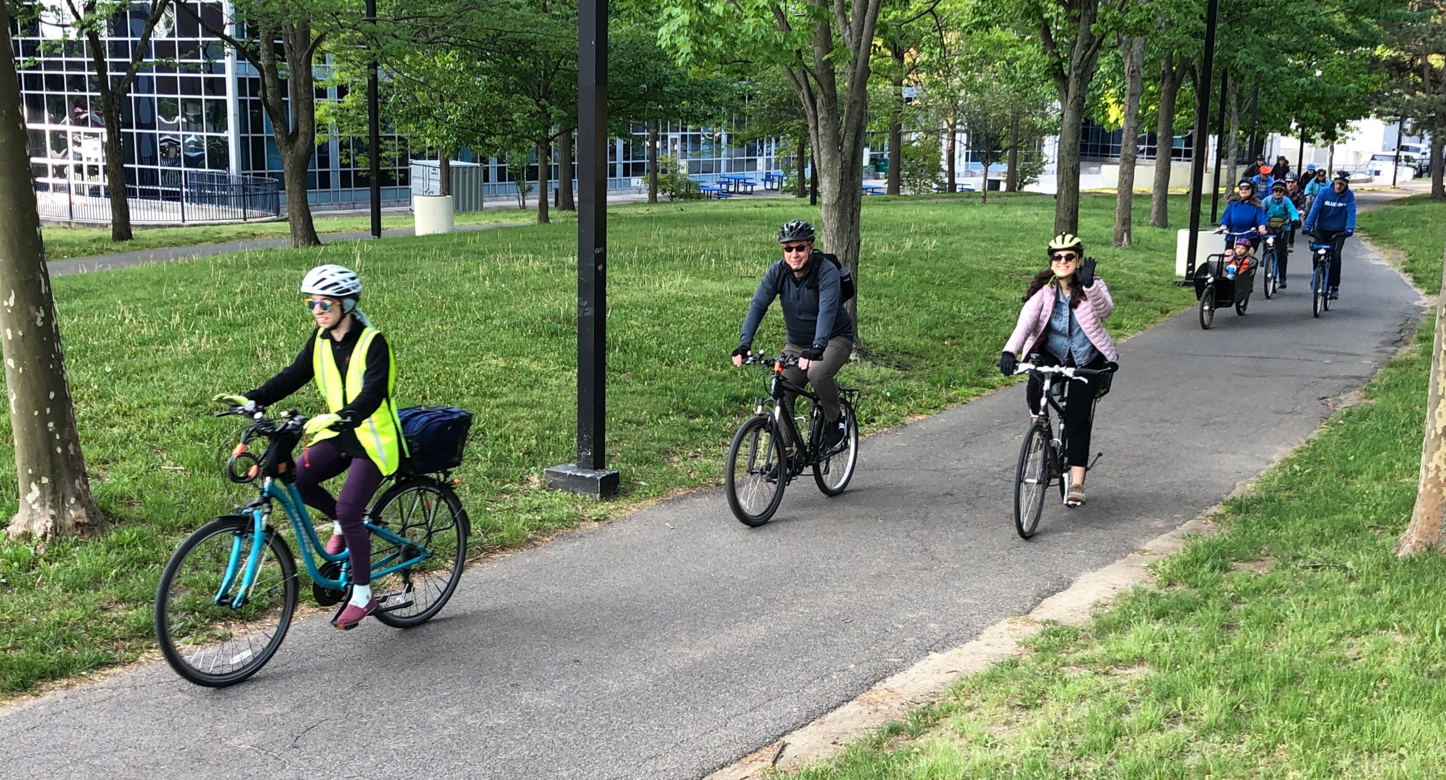 Three people riding bicycles down a paved pathway through a shady park on the SW Corridor. Behind them in the upper right corner is another crowd of people on bikes.