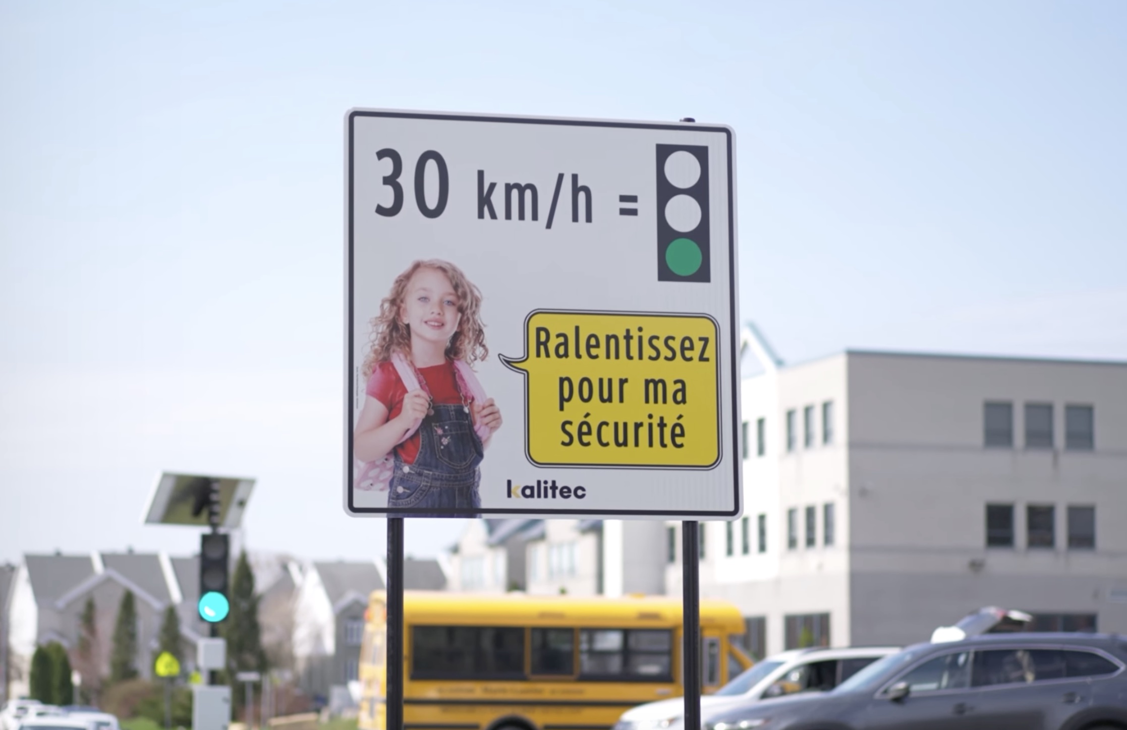 A street sign in French reads "30 km/h" next to a green traffic light; below that, a larger photo of a young girl wearing a backpack is next to a yellow speech bubble that says "ralentissez pour ma securite," or "slow down for my safety." A school bus and a green traffic light are visible behind the sign in the background.