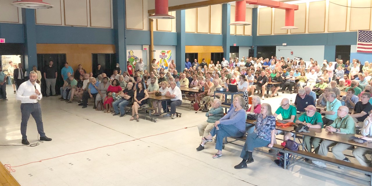 A man in a white collared shirt speaks in front of a large crowd in a school auditorium.