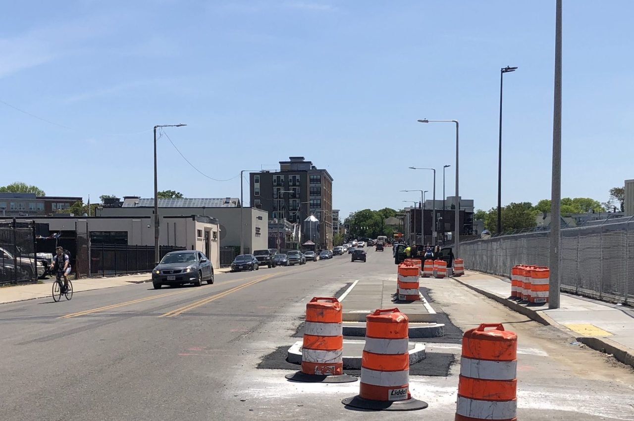 Orange construction barrels surround a newly-constructed median island on a wide roadway in an industrial neighborhood.