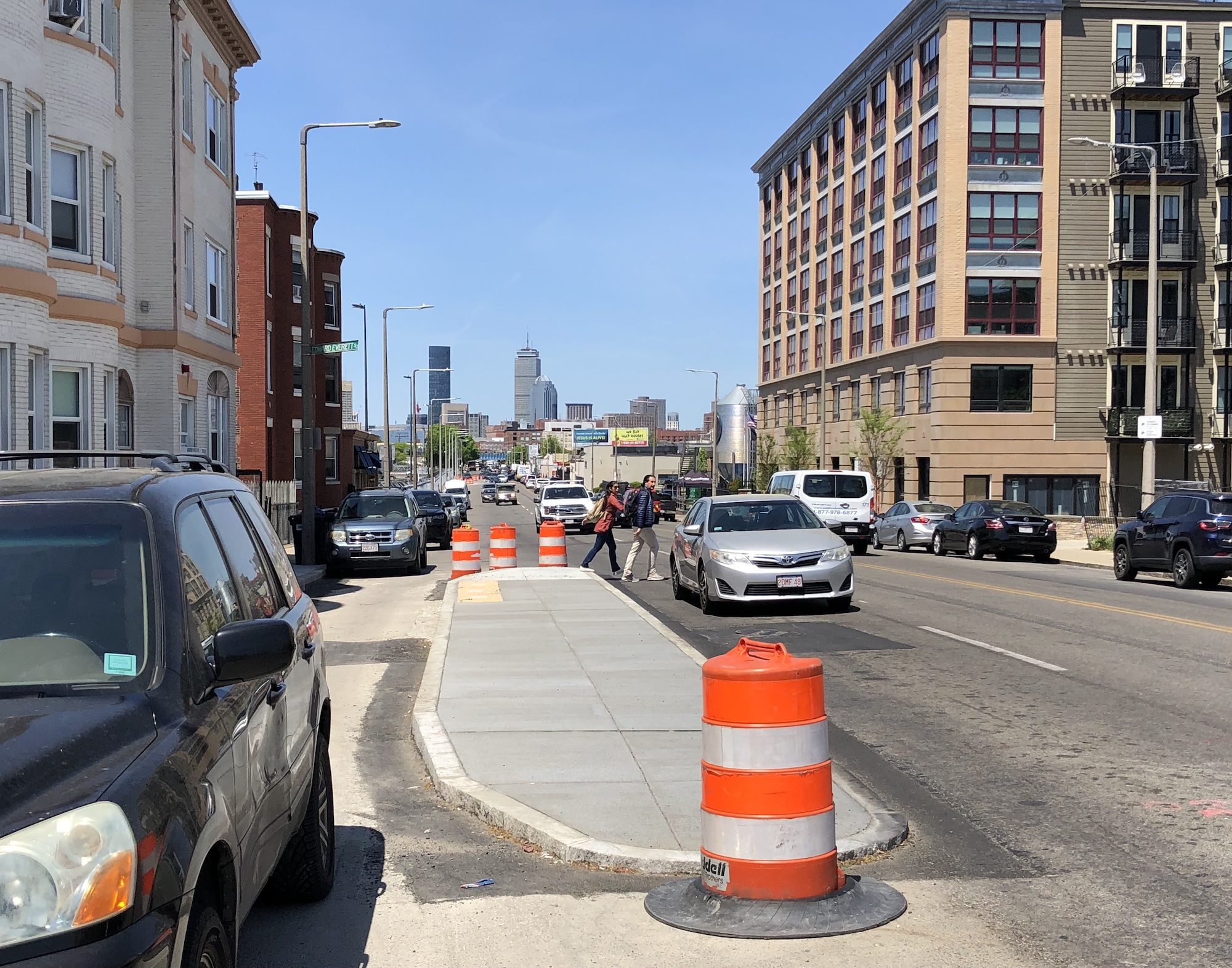 Orange construction barrels surround a newly-constructed median island surrounded by parked cars on the left and moving vehicles to the right. Two pedestrians are stepping off the island to cross the street, which is lined with multi-story apartment buildings. In the distance on the horizon are visible the Prudential Center and other skyscrapers in Back Bay.
