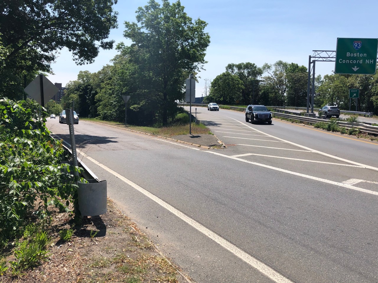 Multiple motor vehicle lanes merge together on a highway surrounded by trees. A green interstate highway sign above the lanes to the right reads "Interstate 93 Boston Concord NH" above a smaller "Exit" sign. 