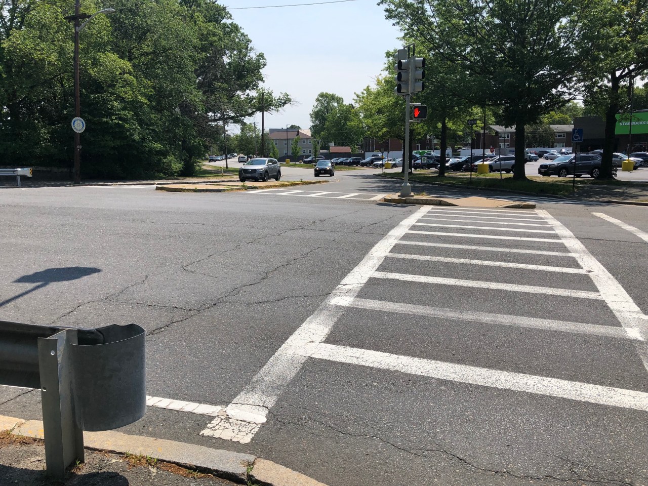 A wide asphalt intersection, with a guardrail in the foreground and a ladder-style crosswalk leading to a traffic island in the distance. In the background, the roadway is lined with leafy trees.