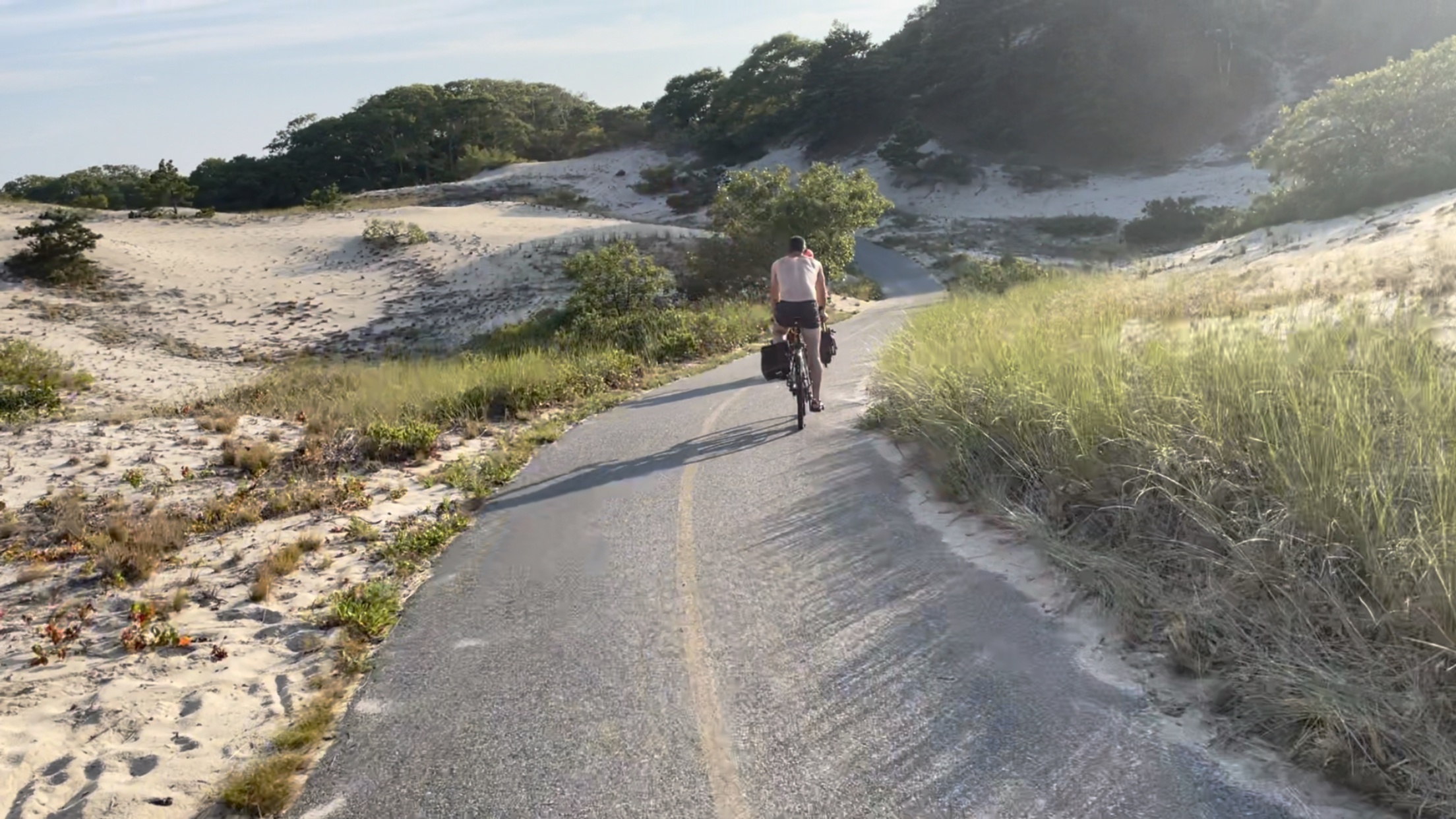 A paved path running through sand dunes. Two people in bikes are in the middle distance.