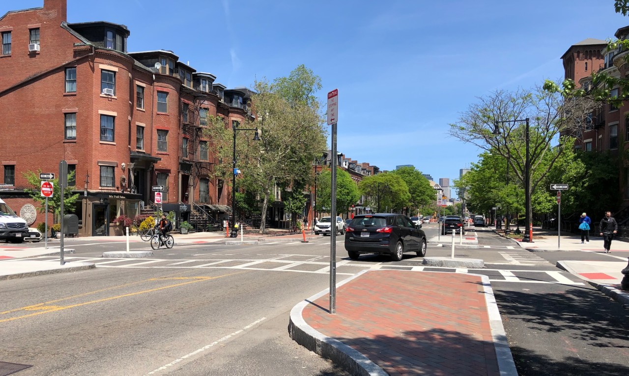 Tremont Street in the South End. In the right foreground is a newly-installed median island, about the size of a parked car, that is paved in bricks and ringed by a granite curb. To its left are two lanes for two-way car traffic; to its right is an asphalt-paved bike lane, and a second curb at the right edge of the photo delineates the street's sidewalk. Historic brick rowhomes line both sides of the street.