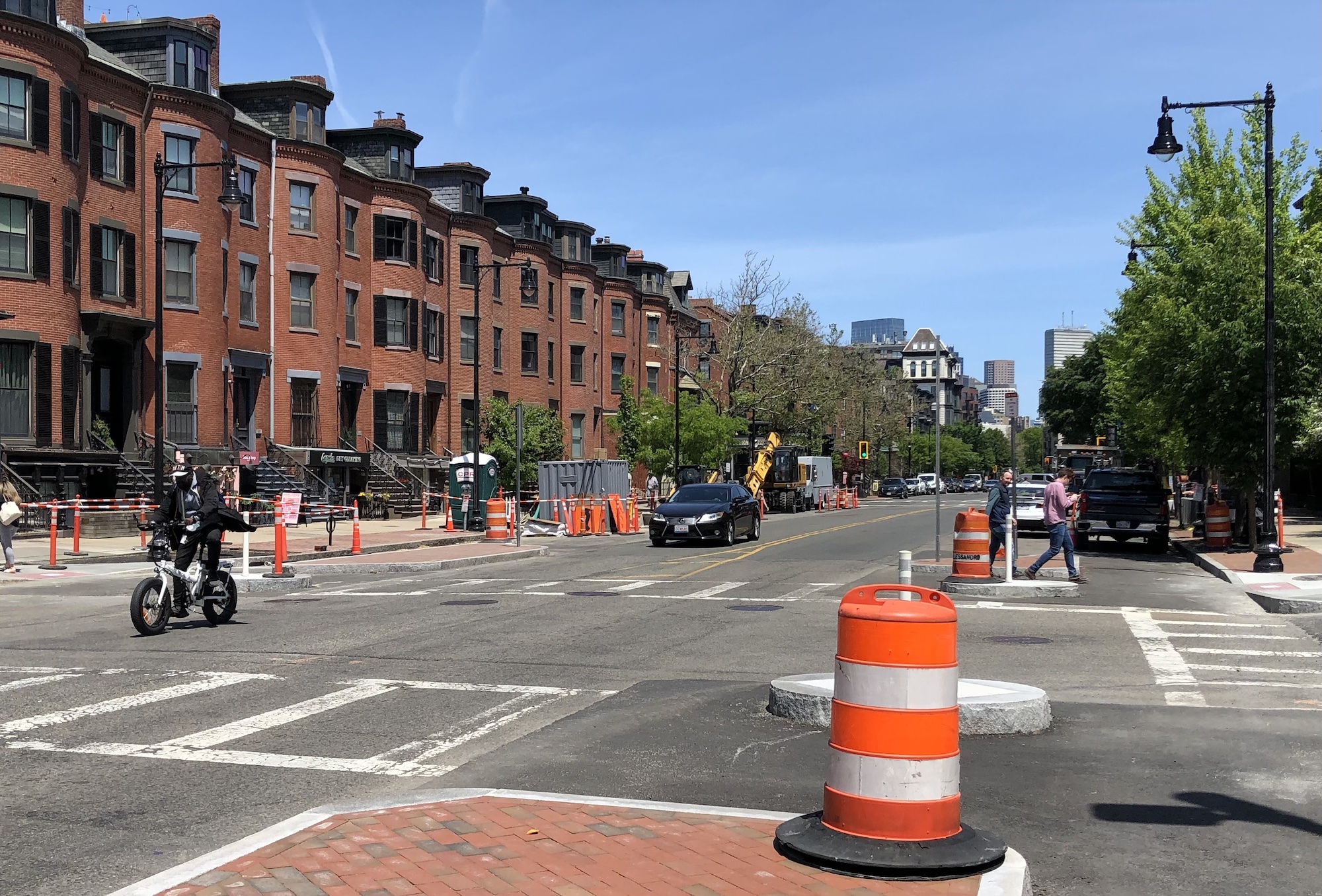 Tremont Street in the South End. In the right foreground is a newly-installed median island paved in bricks and ringed by a granite curb and topped with an orange construction barrel. In the background a pedestrian crosses the street among more construction barrels. On the right, a bicycle rider passes in the opposite lane. Historic brick rowhomes line both sides of the street.