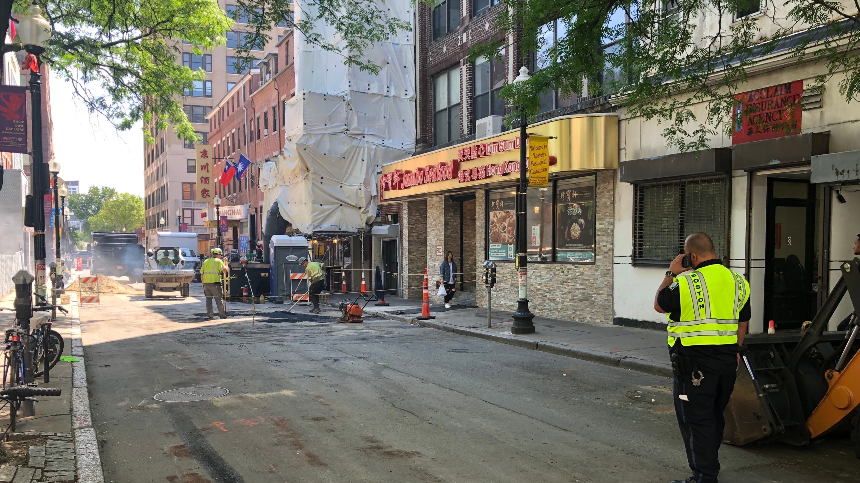 A photo of a street in Chinatown. In the distance on the left, several construction workers shovel asphalt to patch a hole in the street, which is roped off around the construction site. On the right, a Boston Police officer in a green high-viz vest talks on his phone while watching the work.
