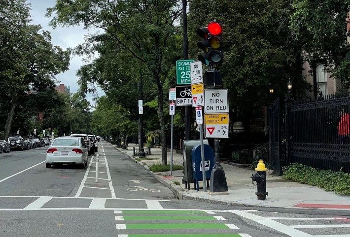 A photo of a traffic light pole in Back Bay in Boston. Two pairs of signs - one facing the camera, the other facing traffic coming from the left - say "no turn on red" and "Turning vehicles yield to [bike and pedestrian pictograms]." Another pair of signs mounted to another pole just beyond the traffic light say "bike lane" and "signals set for 25 mph". The adjacent street has a protected bike lane located between a row of parked cars and the sidewalk.