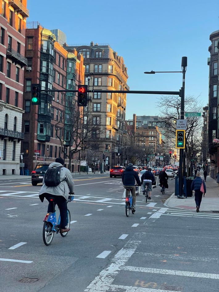 People on bikes wearing winter clothes ride in a bike lane through an intersection in Boston with historic high-rise buildings in the background. A traffic signal above them shows a red right-turn arrow alongside a green bike signal. Signs mounted on the signal pole identify the street as Beacon Street, and another sign says "no turn on red."
