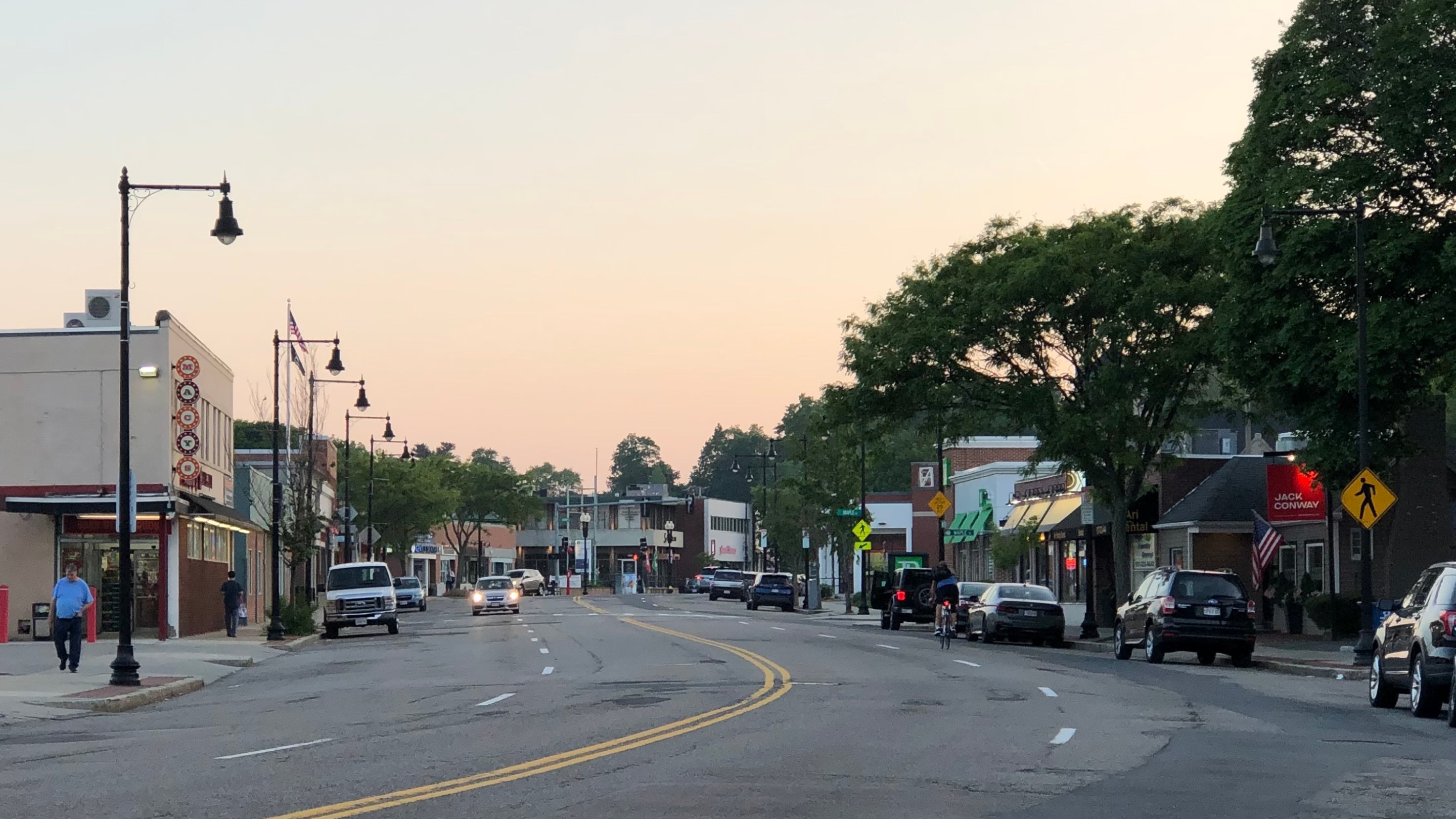 A sunset view down a mostly-empty, wide four-lane street lined with small businesses and parked cars.