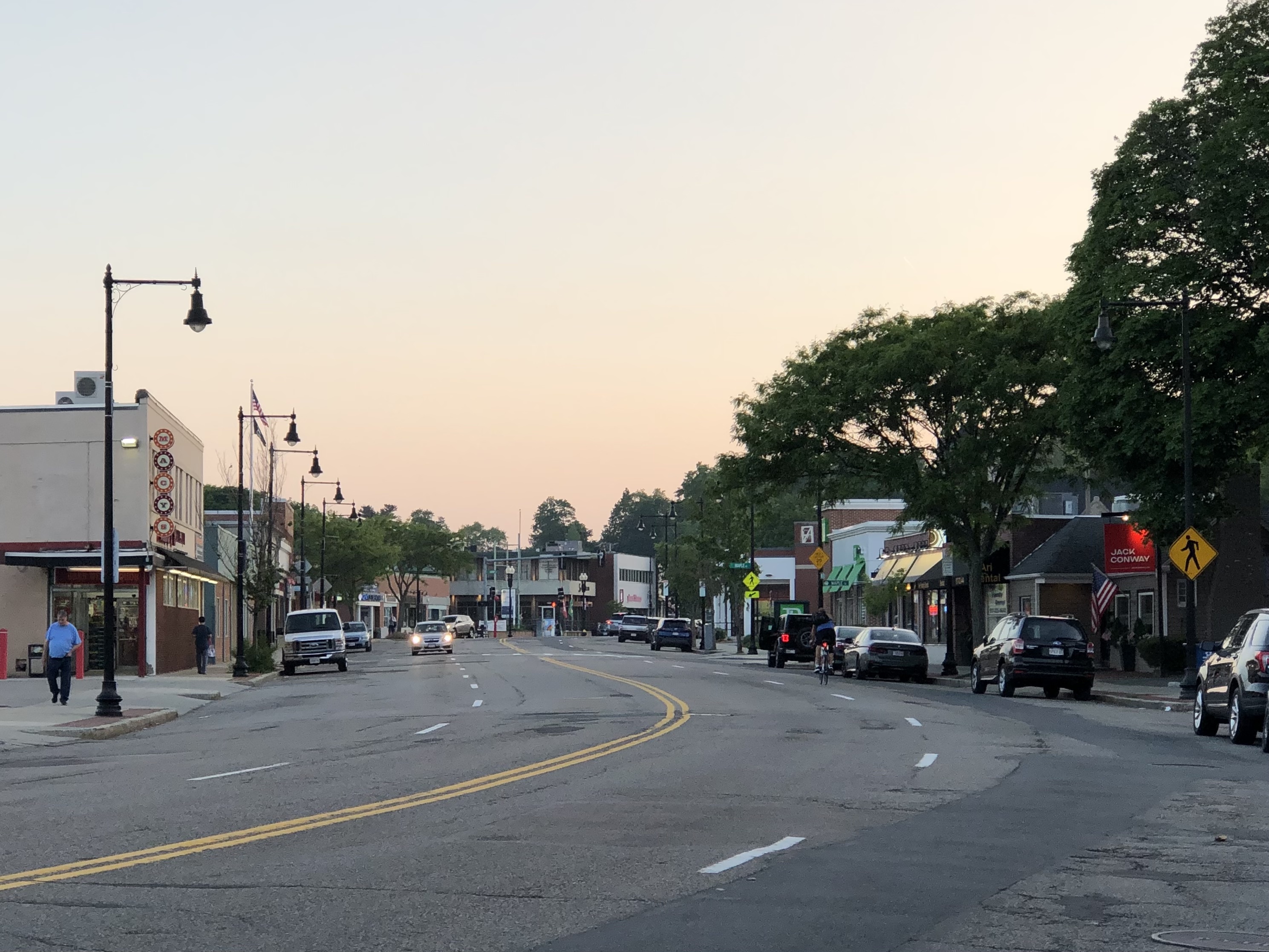 A sunset view down a mostly-empty, wide four-lane street lined with small businesses and parked cars.