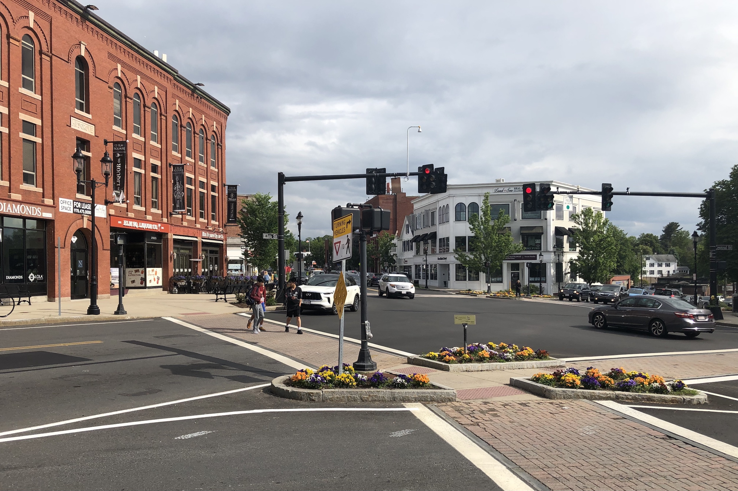 A wide intersection with a small island planted with flowers in the foreground. The street is lined with multi-story buildings with small businesses on the ground floor. A group of three teens crosses the crosswalk in the middle distance.