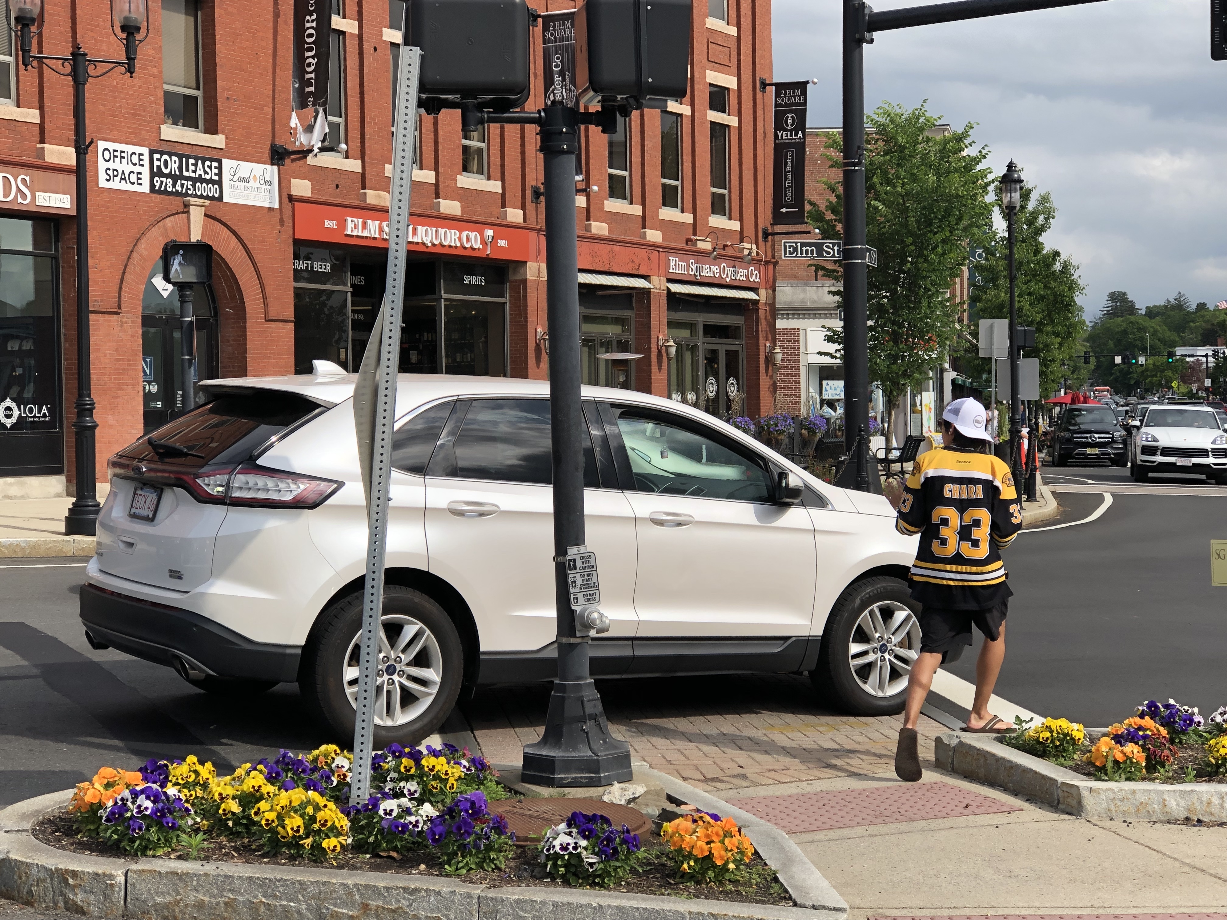 A white SUV blocks a crosswalk while a pedestrian steps up onto the adjacent curb to try and get by. On the other side of the crosswalk a "walk" symbol is visible on the pedestrian signal. Beyond are several small businesses in a brick multi-story building.