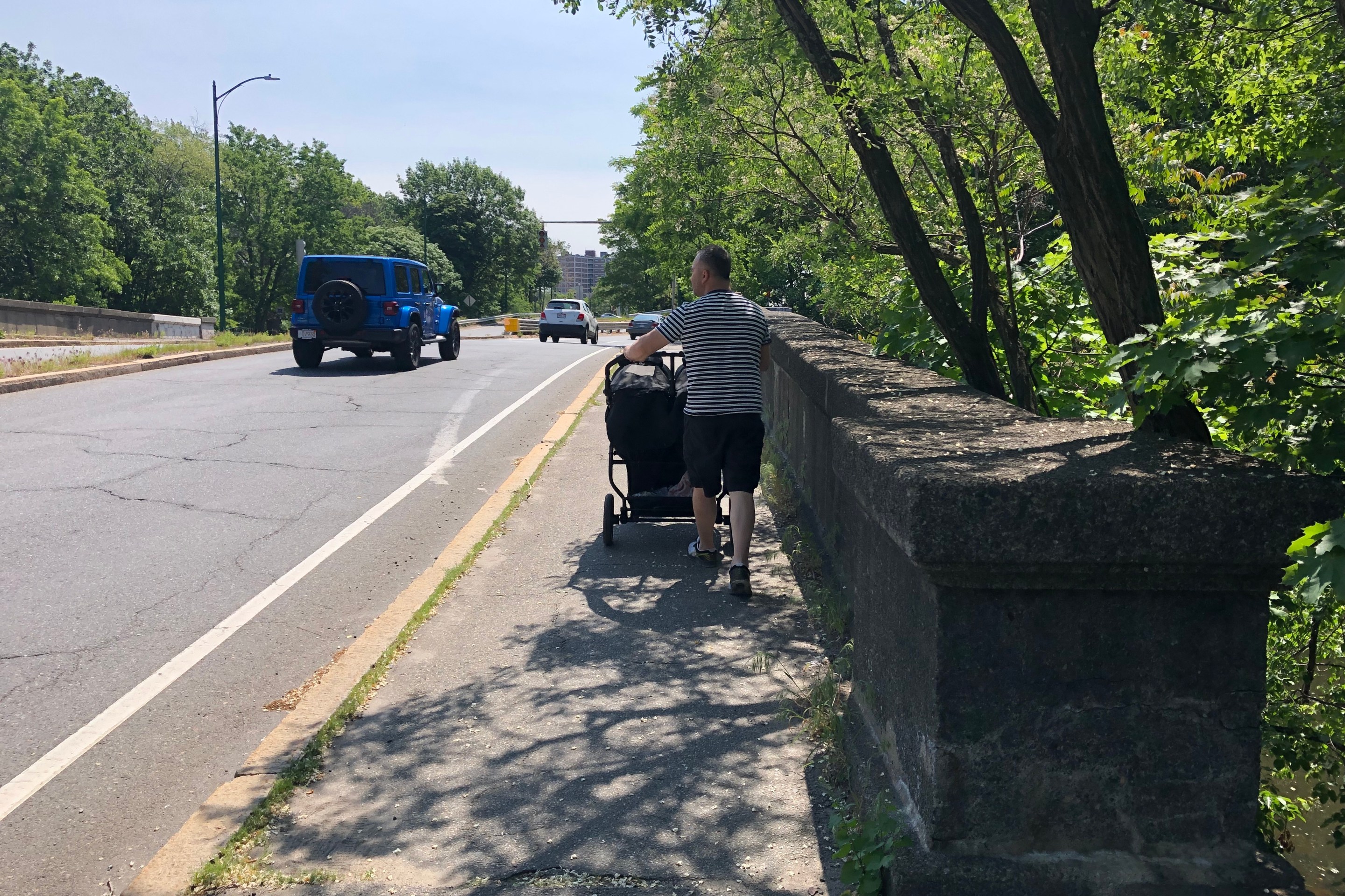 A man pushing a stroller along a narrow sidewalk on a bridge with an old stone masonry railing. Next to him is a wide asphalt highway lane where a large blue pickup truck is driving. In the distance is a brick-clad apartment tower peeking above leafy trees.