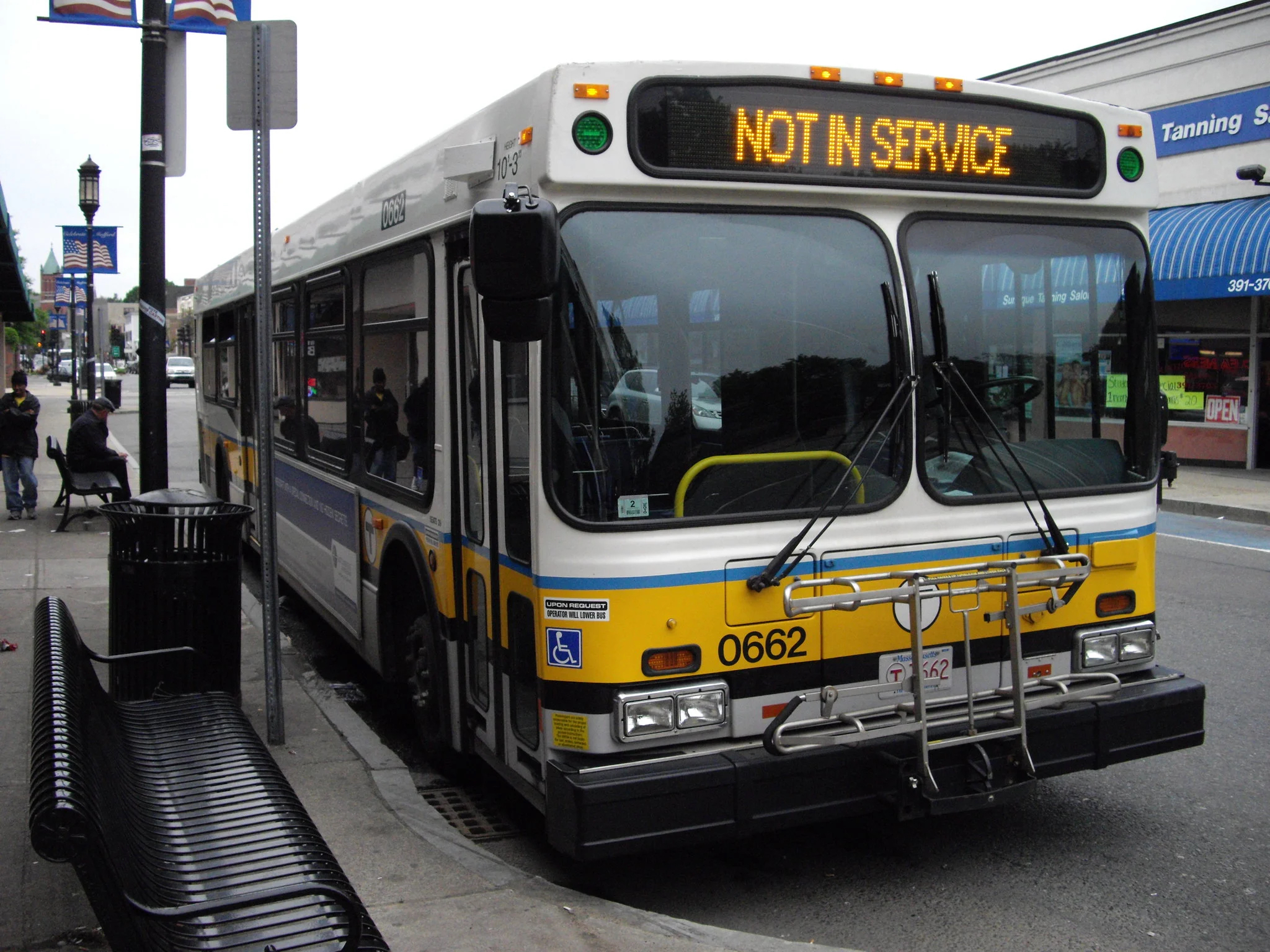 A yellow, white, and black MBTA bus is parked at a curb with the words "NOT IN SERVICE" above the driver's window in the front.