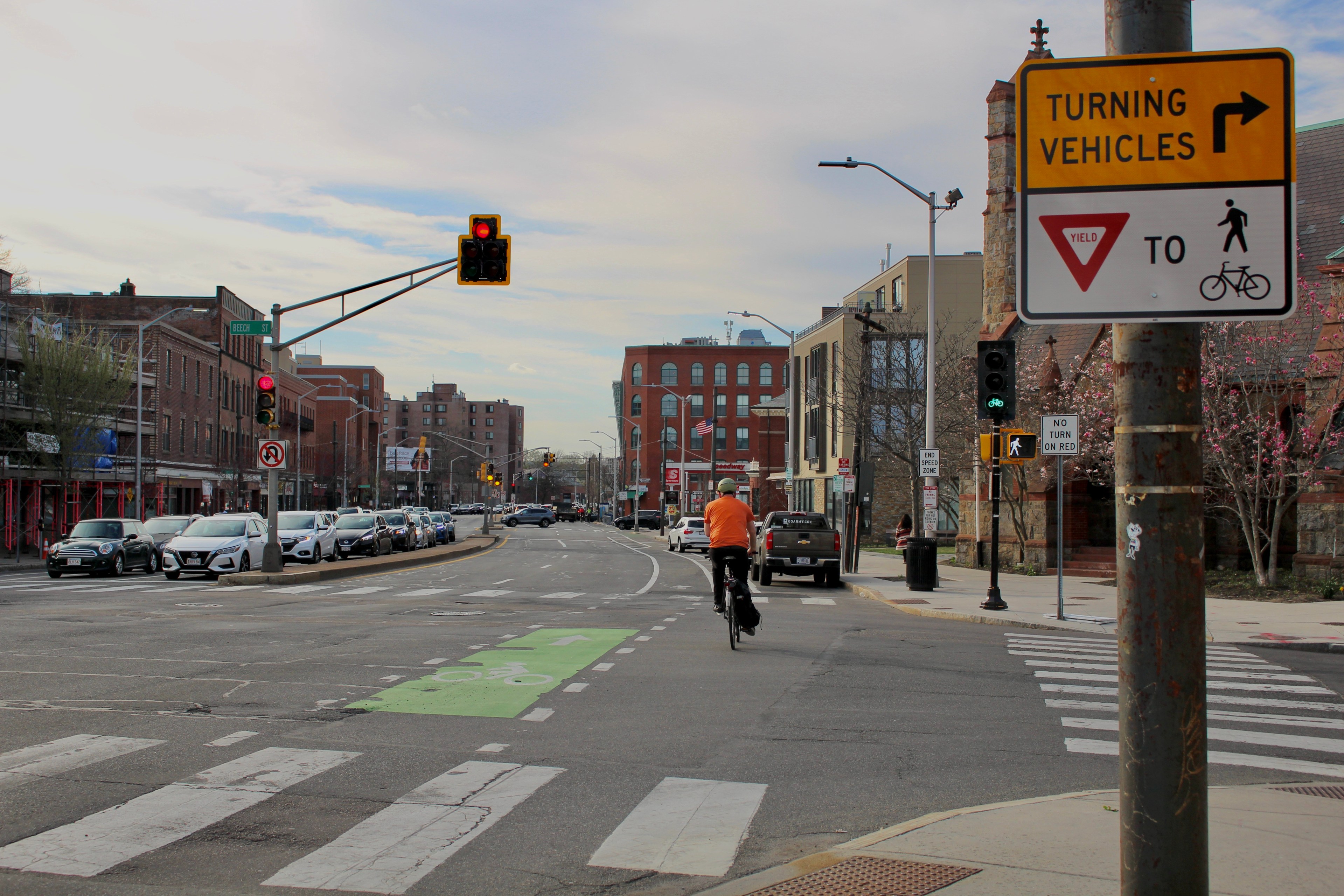 A person on a bike rides past a green bike signal alongside a walk sign while roughly a dozen cars wait at a red light on the opposite side of the street. A sign in the foreground reads "Turning vehicles yield to [bike and pedestrian pictograms]" while a sign next to the walk signal says "No Turn On Red." The street is lined with 3-5 story brick buildings.
