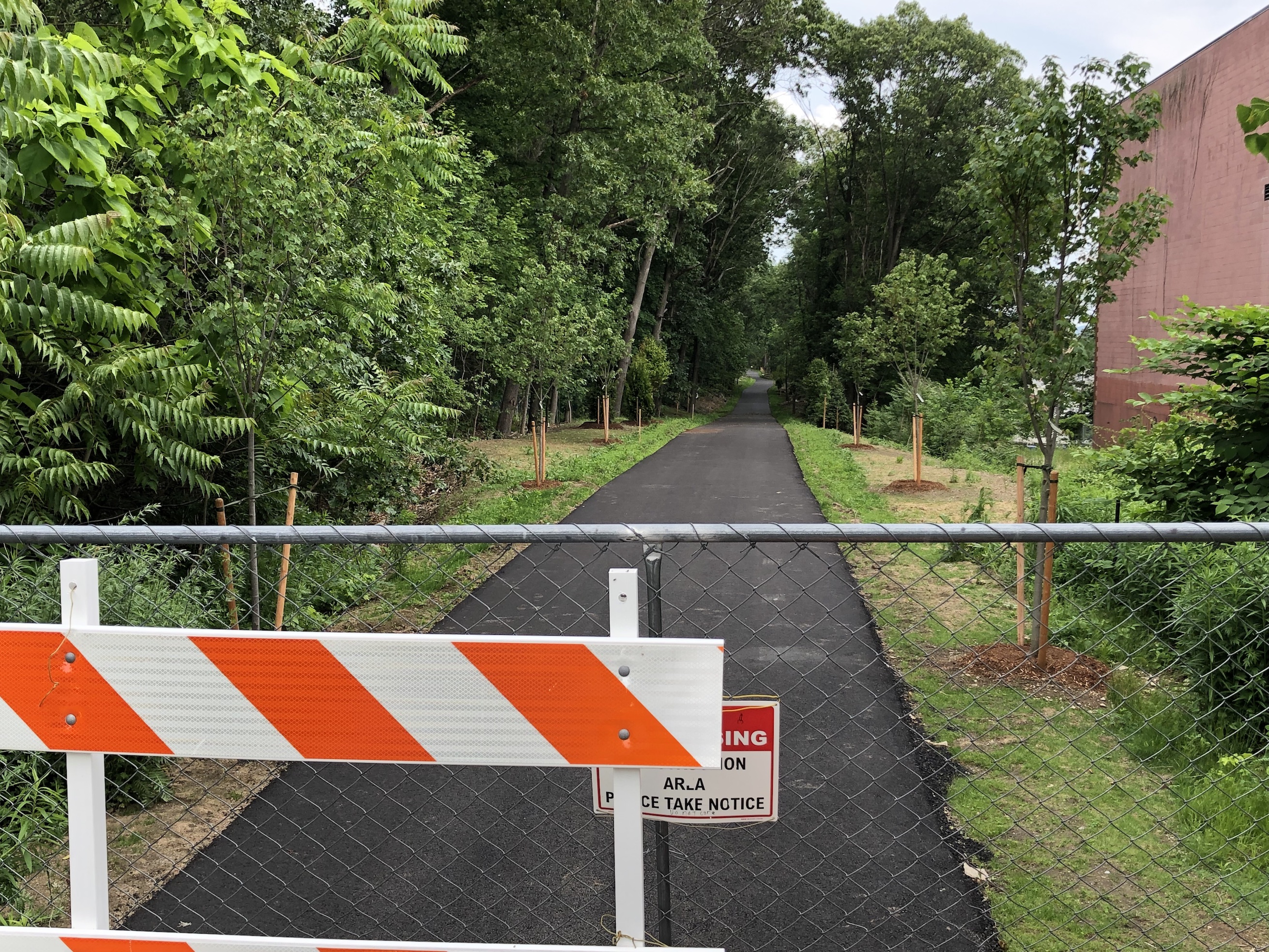 Fencing blocks a newly-paved trail through the woods. On the right is a tan-colored wall with no windows; on the right is a dense grove of trees. The trail disappears into the shade of a tunnel of trees in the distance.