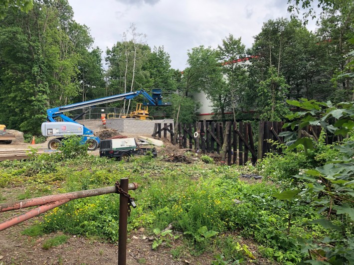 A construction site in a wooded area with meadow plants in the foreground. In the middle are clusters of wooden logs, the remains of an old railroad trestle. To the left is a newly-built bridge abutment made from concrete blocks. A small bulldozer sits atop the abutment next to chain-link fencing. Other construction equipment sits idle to the left. Beyond the trestle is the large windowless wall of a warehouse building.