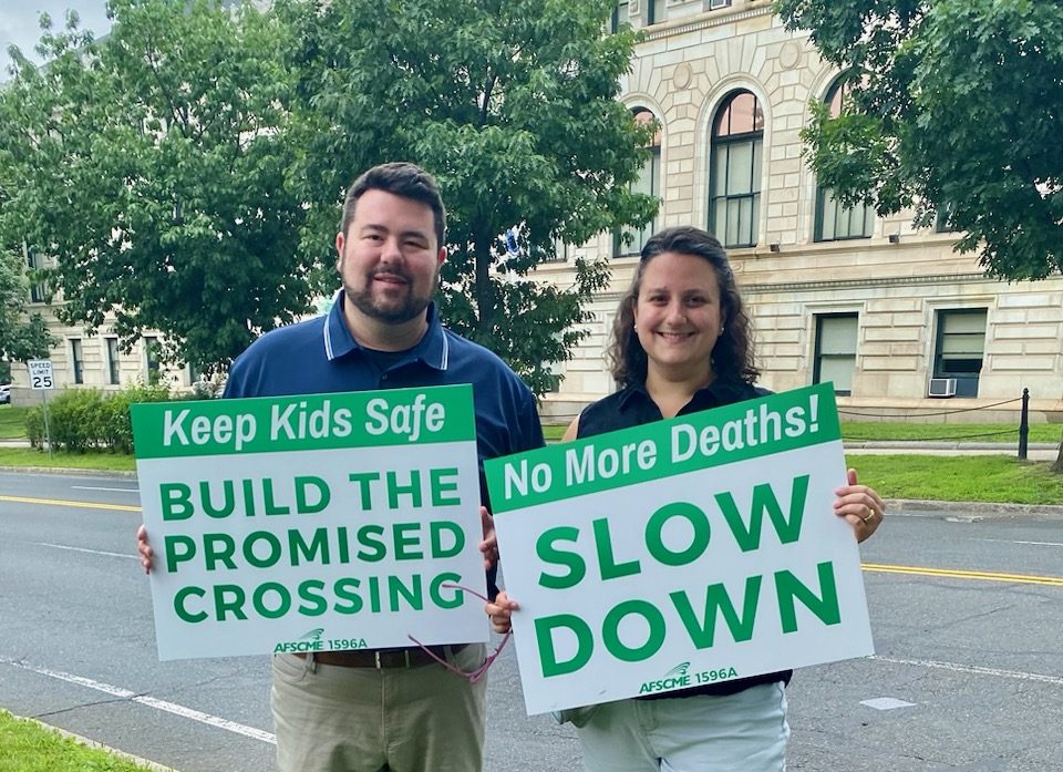 A man and a woman in casual clothing hold signs next to a wide four-lane street. A grand Italianate-style library building with tall arched windows is in the background behind them on the other side of the street. The man holds a sign reading "keep kids safe: build the promised crossing" and the woman holds a sign reading "No more deaths! SLOW DOWN"
