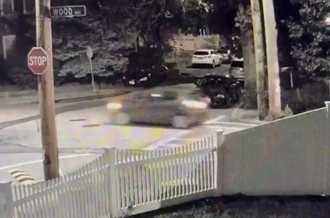A frame from a surveillance video shows a blurry gray hatchback speeding through an intersection at nighttime. In the foreground is a white picket fence.