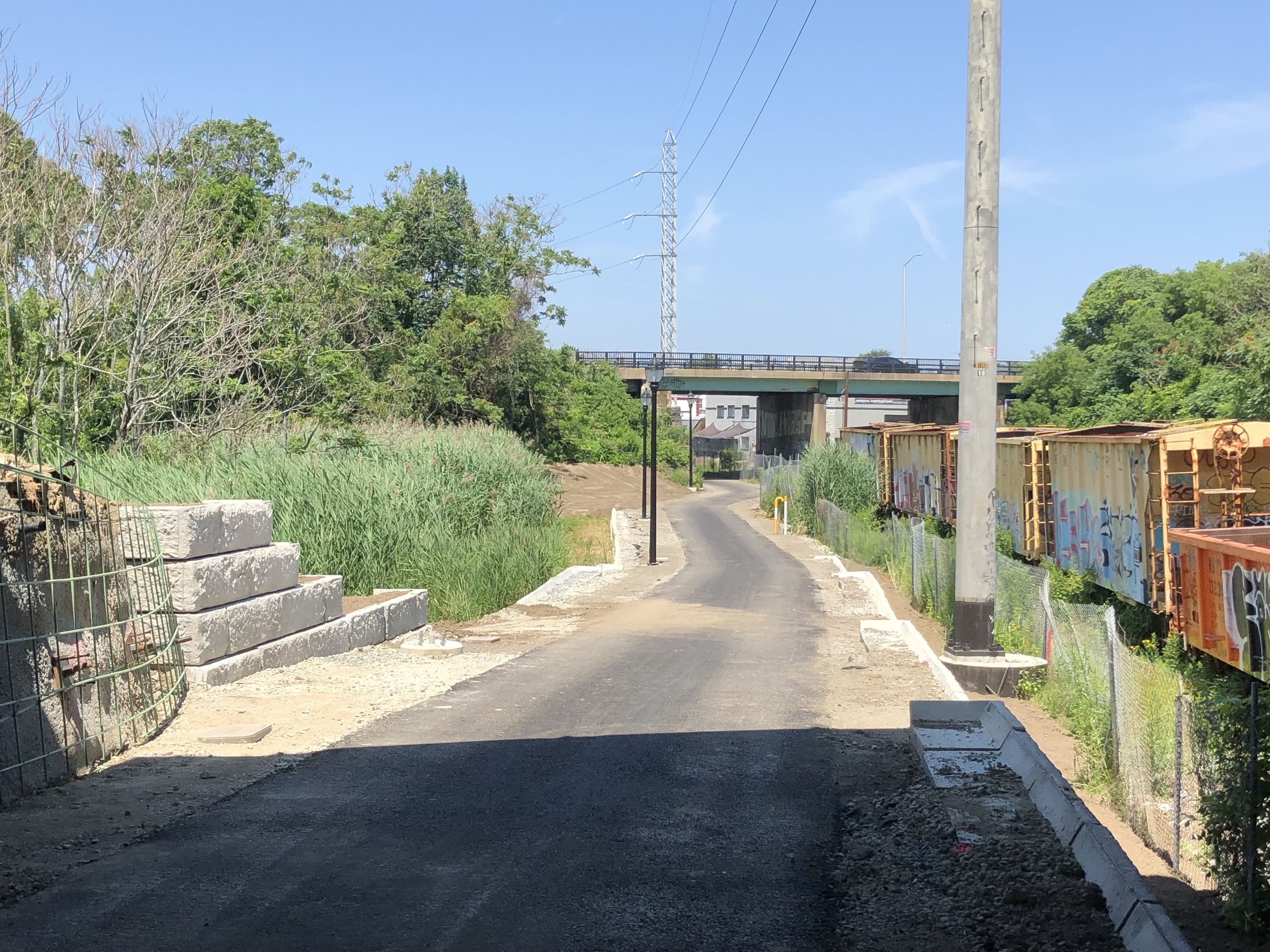 A paved path extends under an overpass in the distance alongside railroad tracks with rusty boxcars and a power line corridor, which run along the right edge of the photo. To the left is a retaining wall and some exposed rebar from an under-construction bridge abutment.