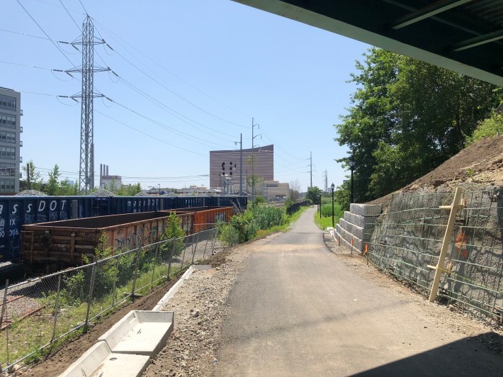 A paved path extends into the distance alongside power lines and railroad tracks, at the left edge of the photo, and an under-construction bridge abutment (right). In the distance is the copper-colored tower of the Encore Casino.