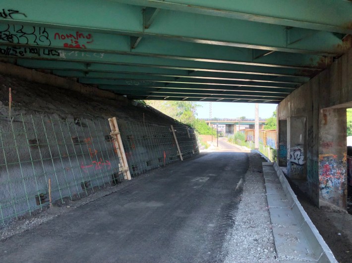 A paved path passes under a highway overpass. To the left is a wall of exposed rebar over a concrete abutment. In the distance, the trail continues under a sunny field and passes under a second highway overpass next to some railroad boxcars.