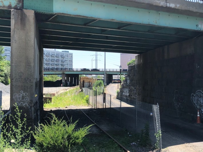 A view under a highway overpass. In the foreground are overgrown railroad tracks; to the right is a chain link fence and a freshly-paved multi-use path, which extends under the bridge and continues under a second bridge in the distance. 