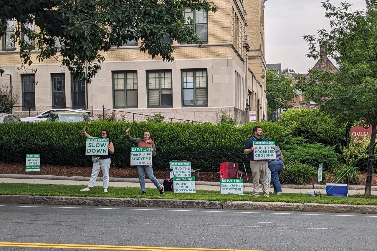 Four people hold green-and-white signs along the curb of a wide four-lane roadway. A grand Italianate-style library building with tall arched windows is in the background behind them on the other side of the street. The man on the right holds a sign reading "keep kids safe: build the promised crossing." One woman to the left holds a sign that reads "Drive like your kid has storytime here" and the woman at the left edge of the photo holds a sign reading "No more deaths! SLOW DOWN." A fourth person on the right is walking towards the sidewalk behind the people holding signs.