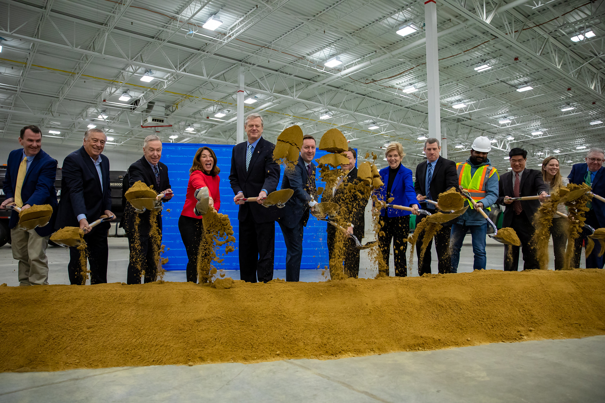A row of men and women, most of whom are wearing business suits, toss shovelfuls of dirt into the air inside an empty warehouse building in front of a blue backdrop that's been set up for a press conference.