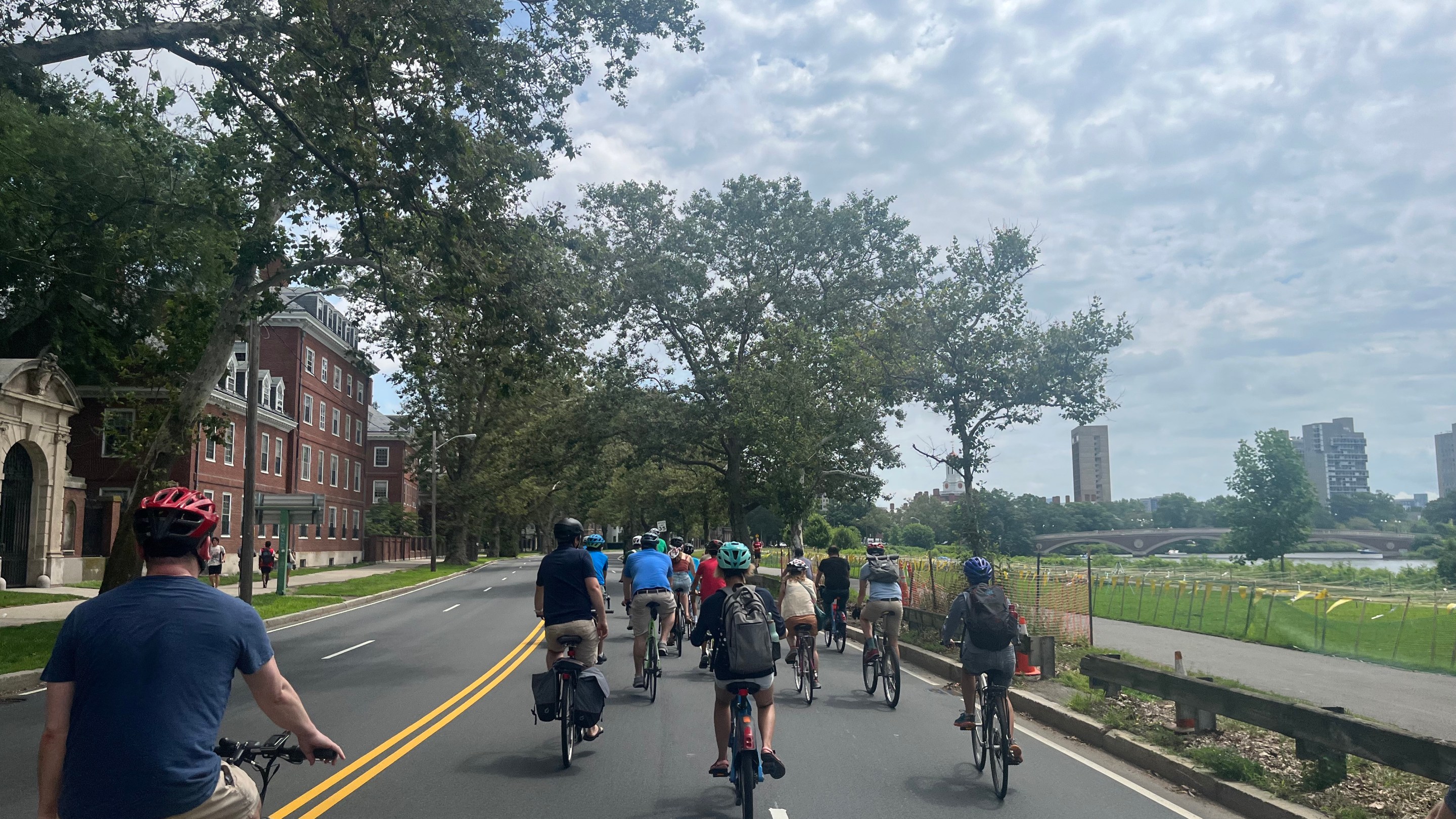 A crowd of people on bikes rides down an empty four-lane highway. On the left side of the roadway are large trees and multi-story brick buildings; on the right is the Charles River with the Boston skyline in the distance.